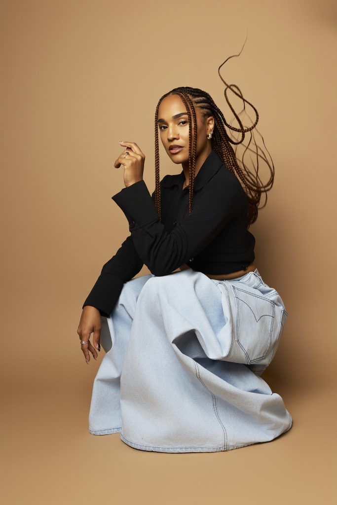 KARABA posing in a black crop top and light wash jeans against a tan backdrop, with hair tossed in the air, reflecting her dynamic energy as a DJ and producer in the Afro-house music scene.