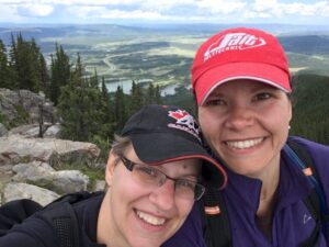 Breanne at left and Jill at right are shown wearing baseball caps and sitting atop a mountain with a valley in the background.