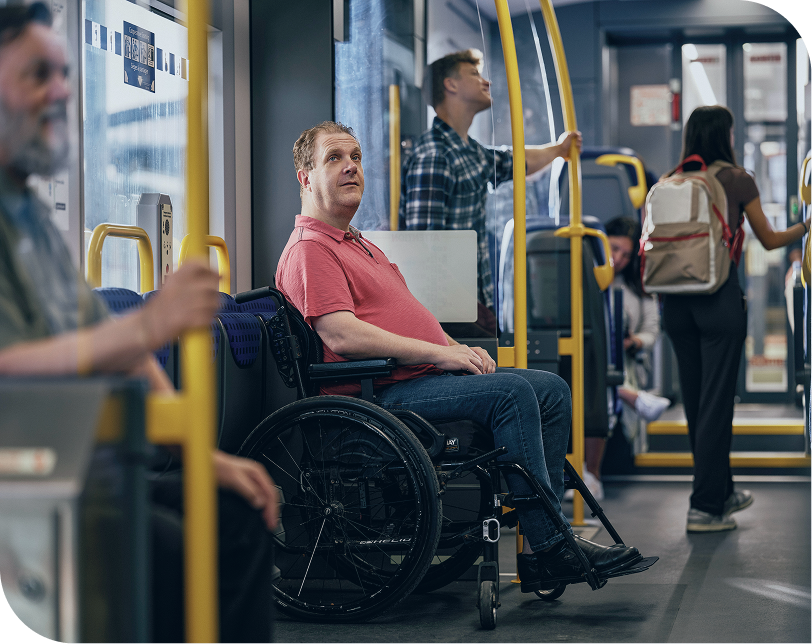Person in a wheelchair inside a bus, with other people standing in the background and foreground.