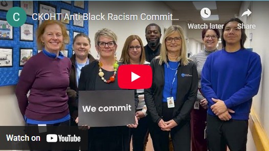 Image of video clip from YouTube. Image shows a group of Healthcare Providers standing in a hallway with one person in the middle holding a sign that says We Commit.