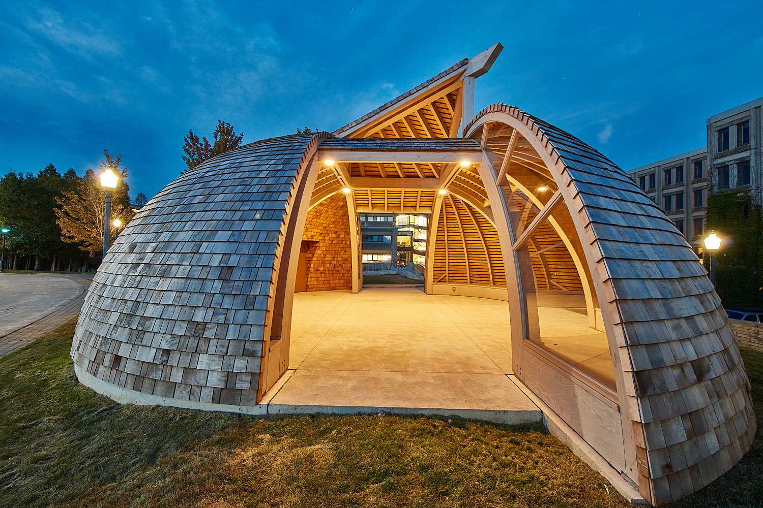 An outdoor gathering space modeled after Anishinaabeg roundhouses, with brown cedar shakes on the sides and open wooden framing inside. 