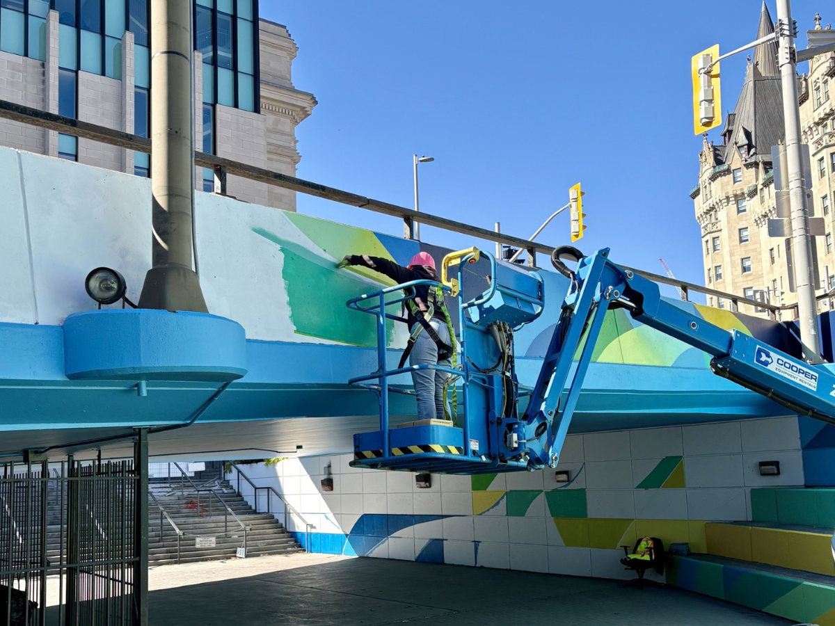 Artiste qui peint une murale composée de diverses formes aux couleurs vives, en bleu, vert et jaune, le long du passage inférieur Rideau-Sussex