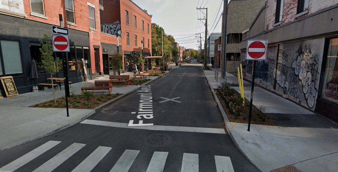 A one lane side street with planted areas and patios next to the sidewalk.