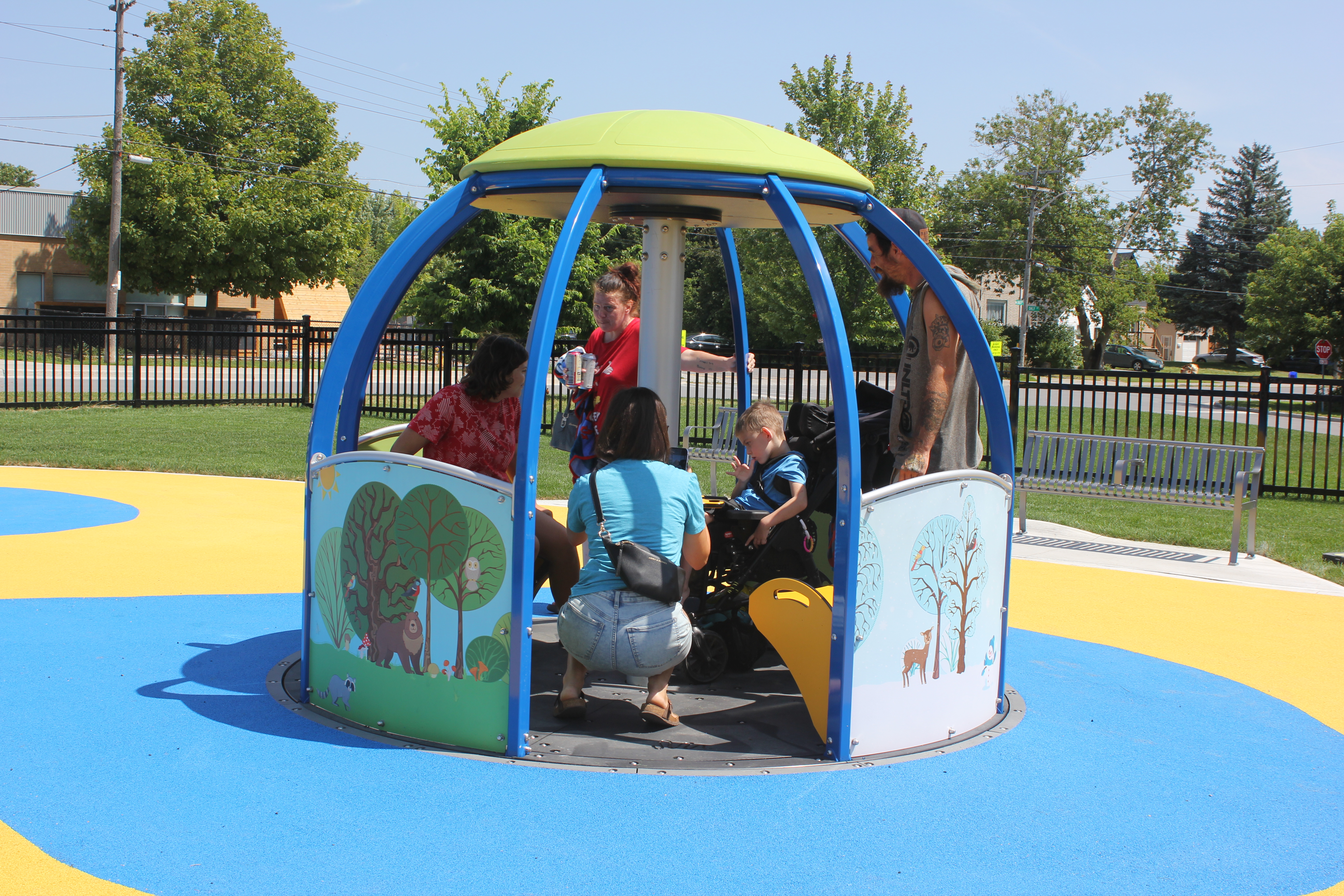 A child in a wheelchair/stroller sits in an accessible spinning play structure. Four adults stand by, one is taking a photo of the child enjoying the experience. 
