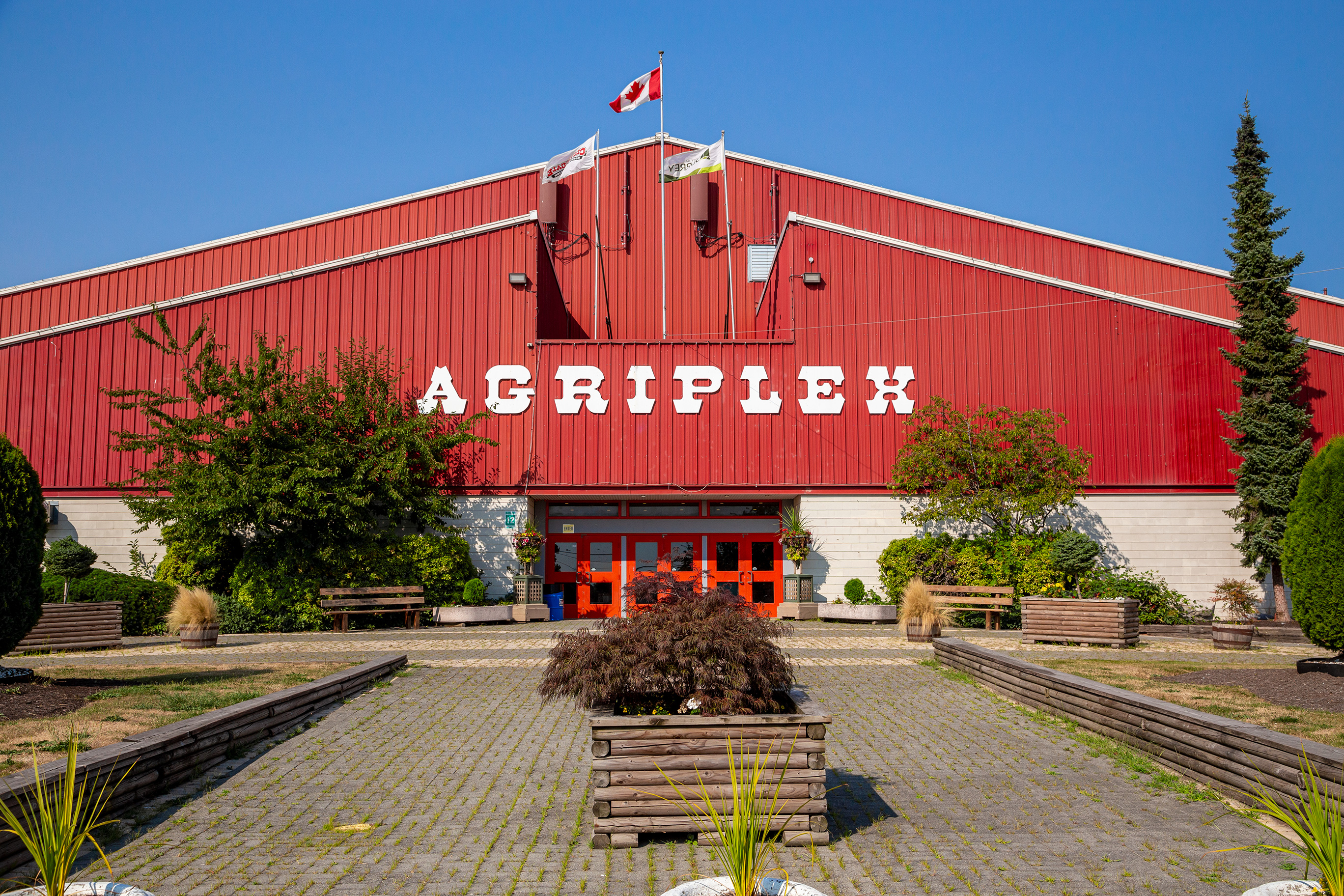 Front entrance of the red Agriplex building, with large white “AGRIPLEX” lettering on the facade, flags flying above the entrance, and landscaped planters lining a paved walkway.