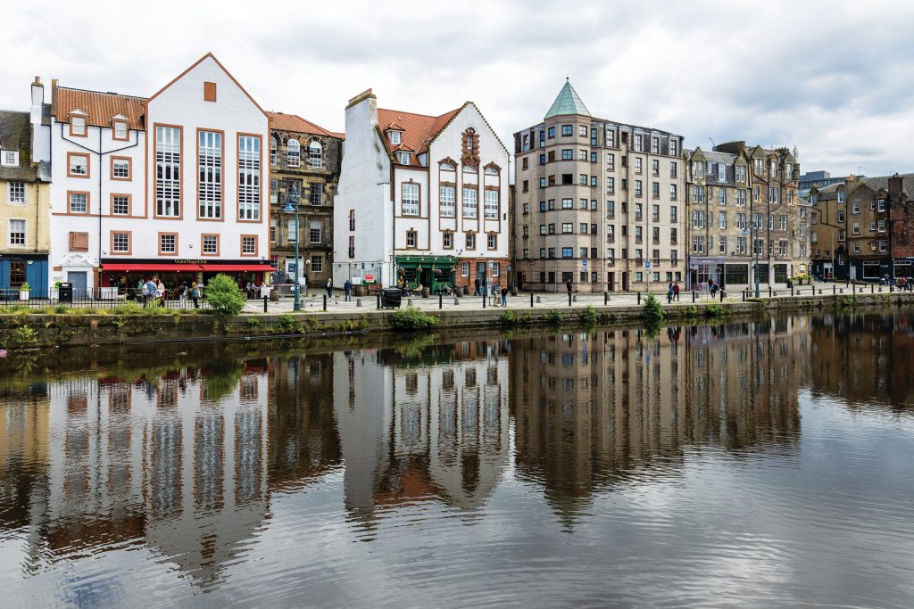 Scottish restaurants, pubs, and boutiques line a waterfront, with buildings reflecting on the water.