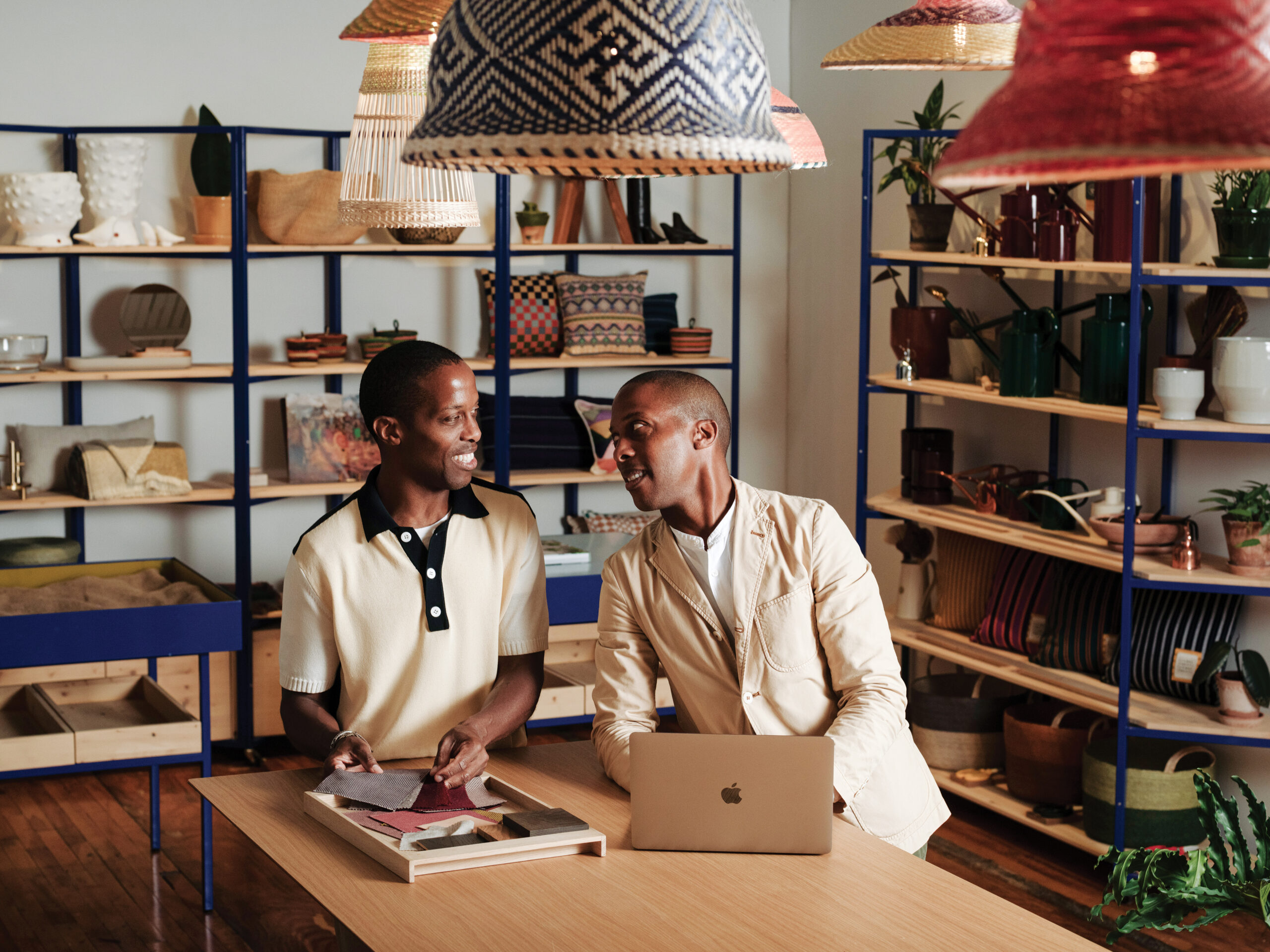 Two men engaging in conversation at a wooden table, surrounded by colorful homeware and decor items on shelves, with a laptop and fabric swatches on the table, reflecting the creative atmosphere of Goodee's office in Montreal.