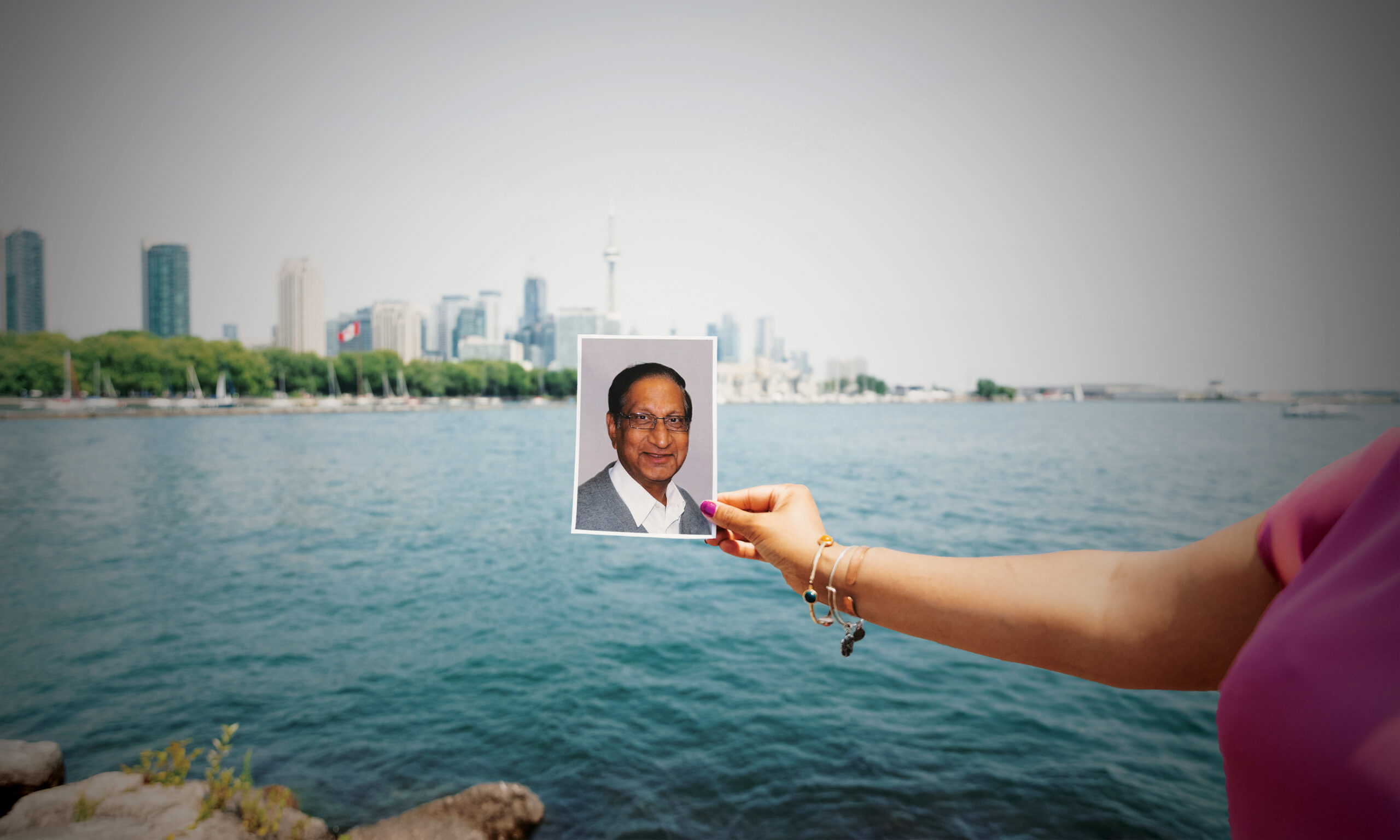 Person holding a photo of an elderly man against a waterfront city skyline, representing themes of memory and cultural connection.