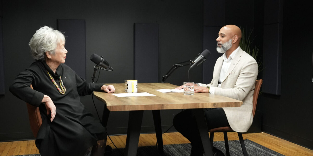 Podcast hosts engaged in conversation at a table, discussing leadership and cultural identity, with microphones and water glasses visible.