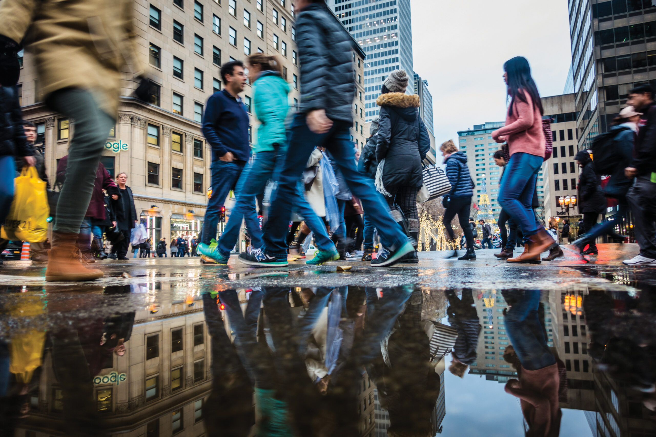 Crowd of pedestrians crossing a city street with reflections in a puddle, showcasing urban life and diversity, relevant to discussions on immigration in Canada.