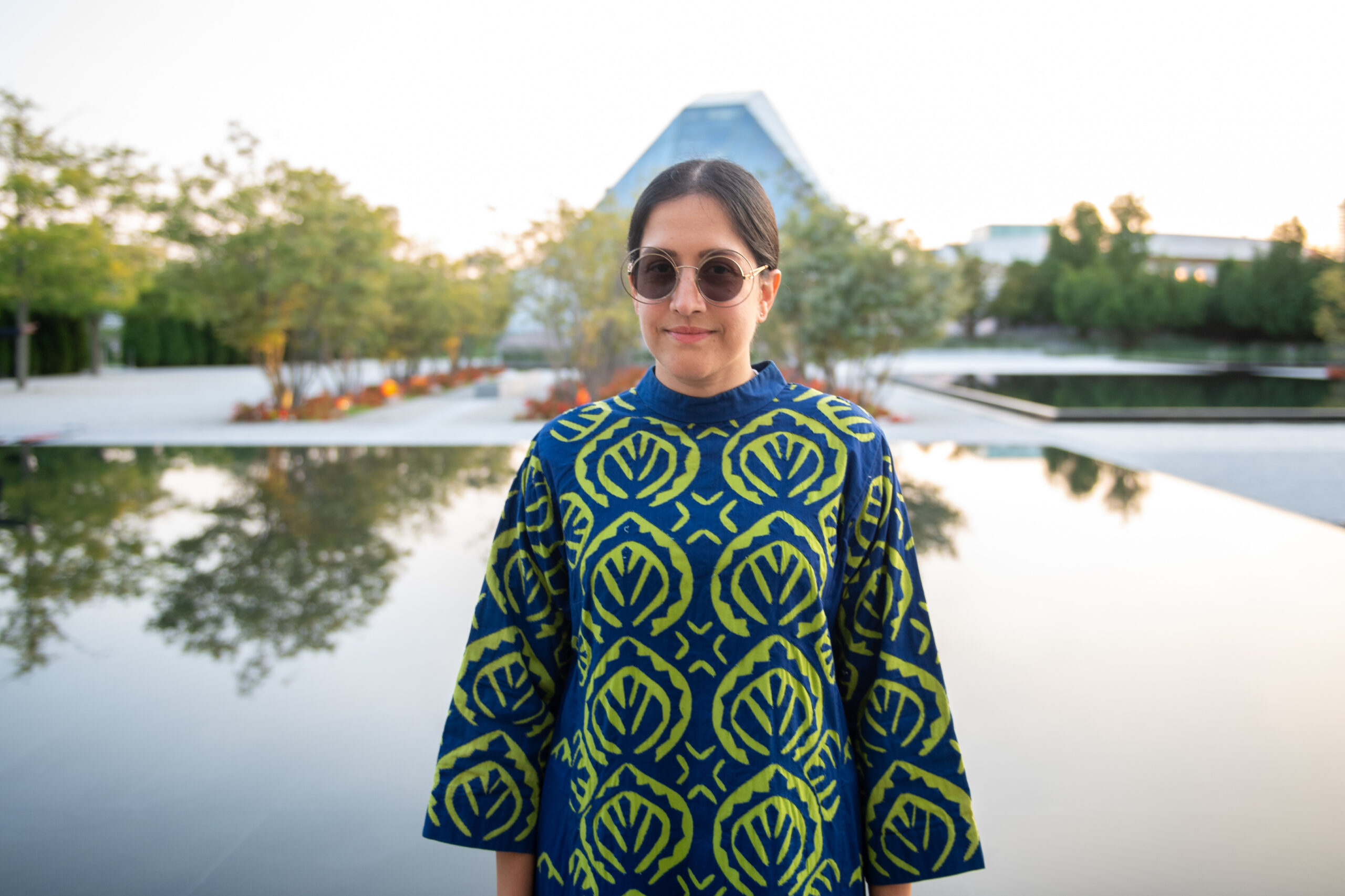 Woman in a vibrant blue and green patterned garment, standing by a reflective water surface surrounded by greenery, evoking a stylish atmosphere in a cultural context.