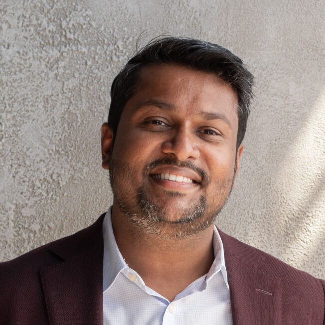 Kumaran Nadesan, Chief Executive Officer of Three Magazine, smiling against a textured wall, representing leadership in cross-cultural storytelling and global identity.