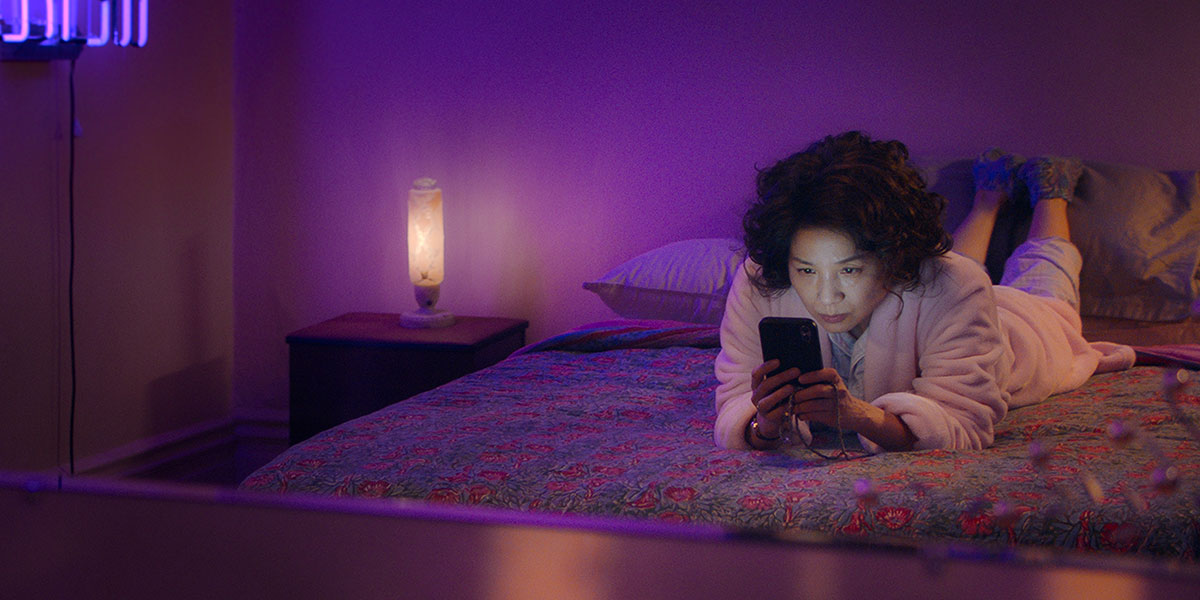 Woman in a pink robe lying on a bed, focused on her smartphone, illuminated by soft purple lighting, reflecting a cozy, modern lifestyle setting.
