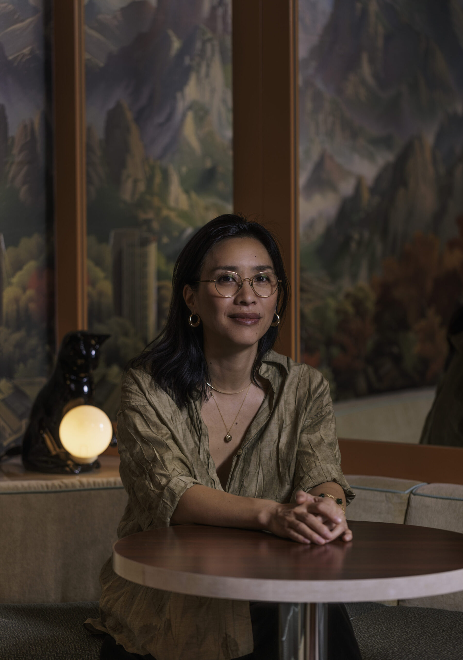 Portrait of Tannis Ling, restaurateur, seated at a table in a modern, artistic caf&eacute; setting, with a black cat sculpture and a scenic mural in the background, reflecting cultural cuisine and lifestyle themes.