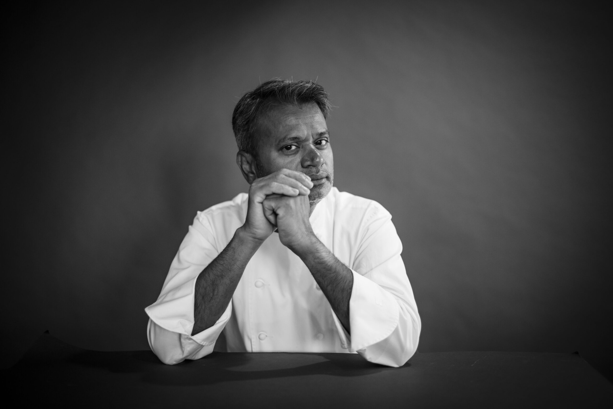 Chef Sash Simpson in a white chef's coat, thoughtfully posing with hands clasped, against a dark background, reflecting his culinary expertise and philanthropic efforts in food.