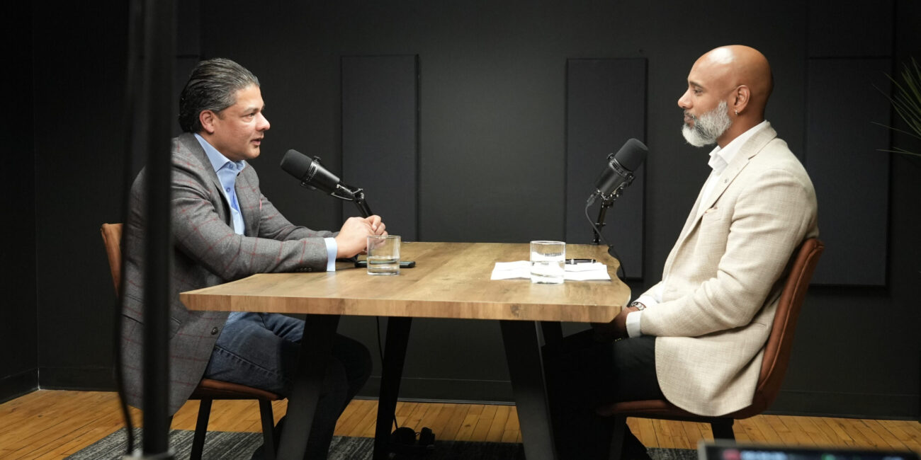 Two men engaged in conversation at a podcast recording table, featuring microphones, glasses of water, and a wooden tabletop, set against a dark backdrop, reflecting themes of leadership and cultural identity.
