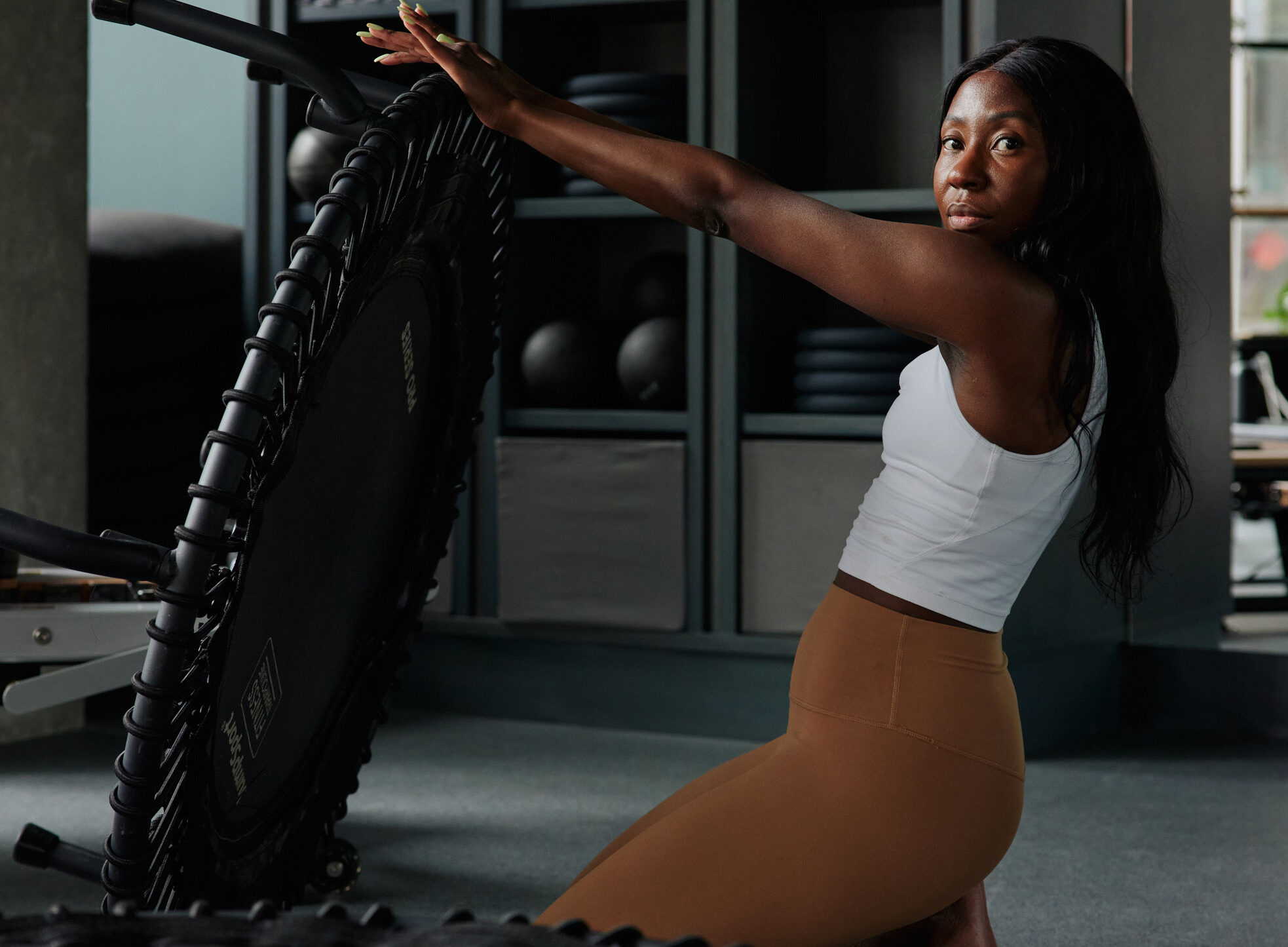 Woman exercising with a trampoline in a modern fitness studio, wearing a white tank top and brown leggings, emphasizing wellness and lifestyle themes.
