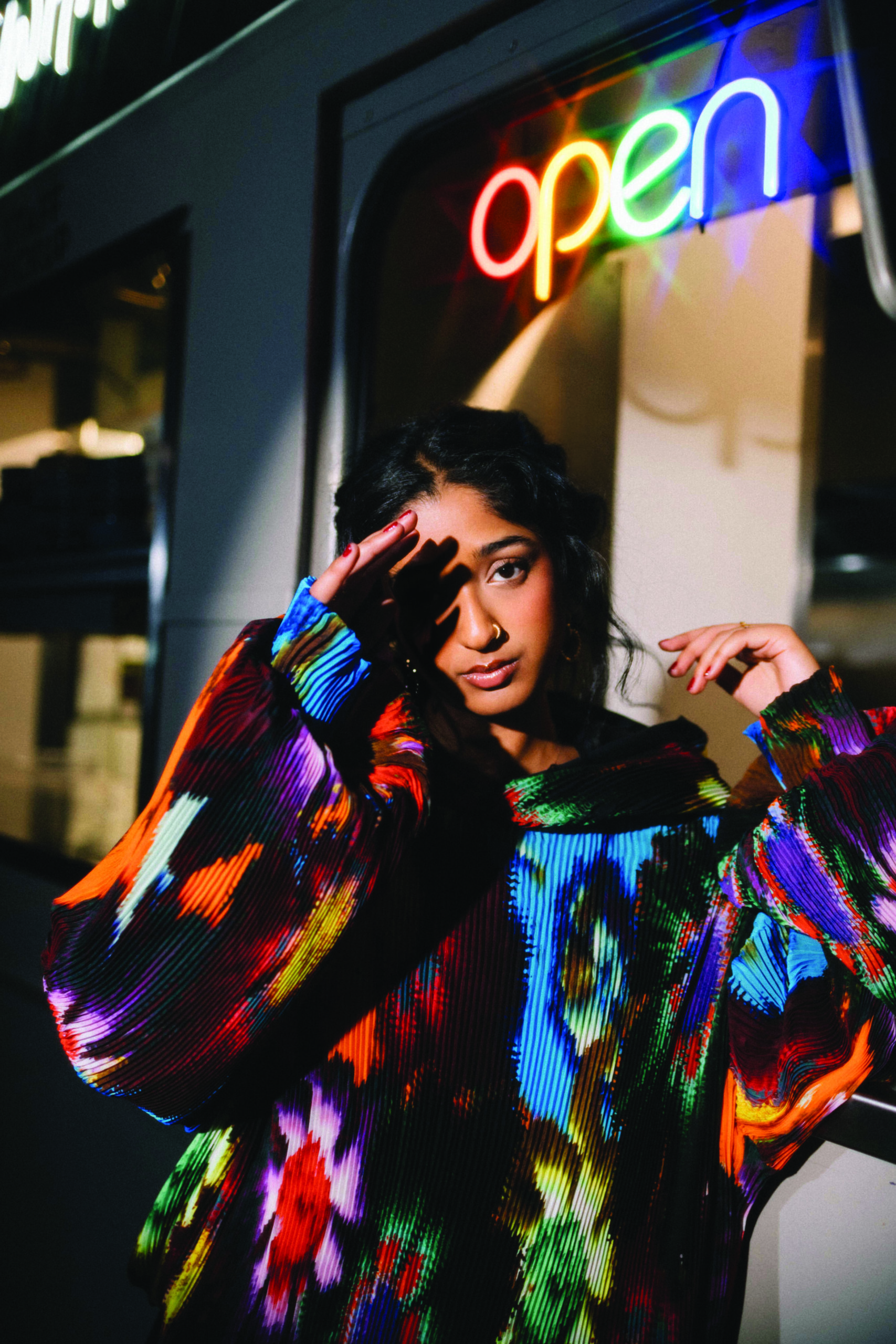 Young woman posing in colorful oversized sweater with vibrant floral patterns, illuminated by neon "open" sign in the background, reflecting the return of bold fashion choices.
