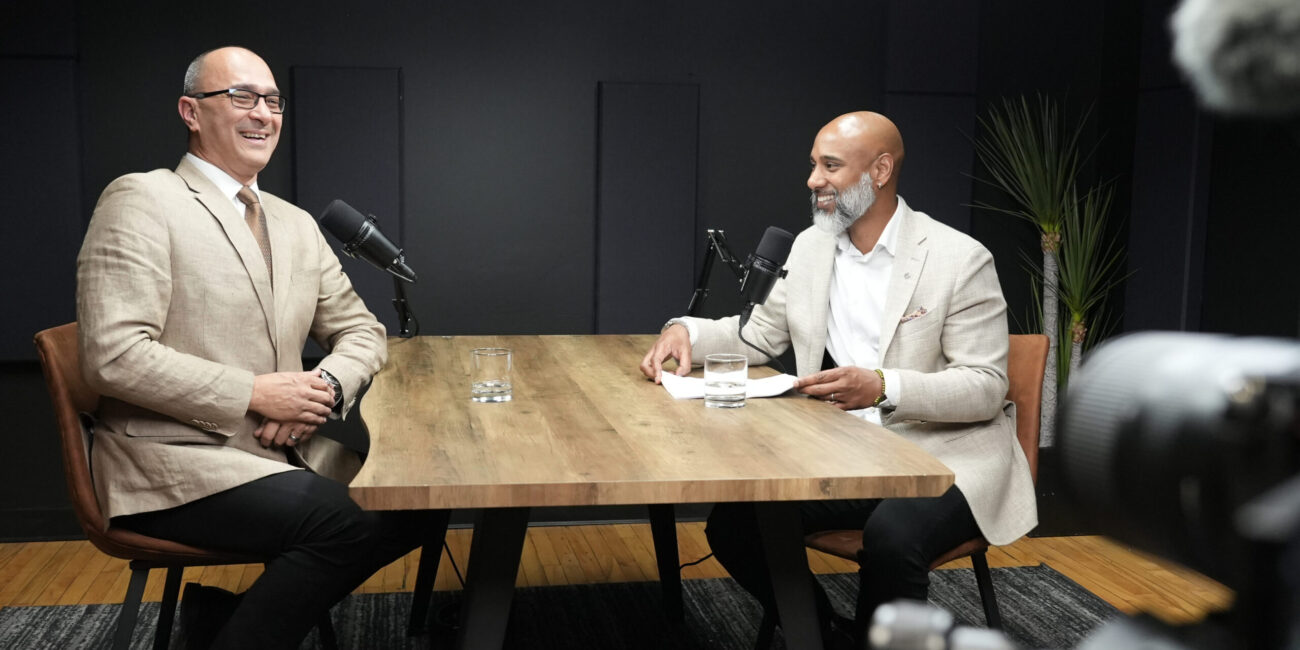 Two men engaging in a podcast discussion at a wooden table, featuring microphones and glasses of water, set in a modern studio environment.