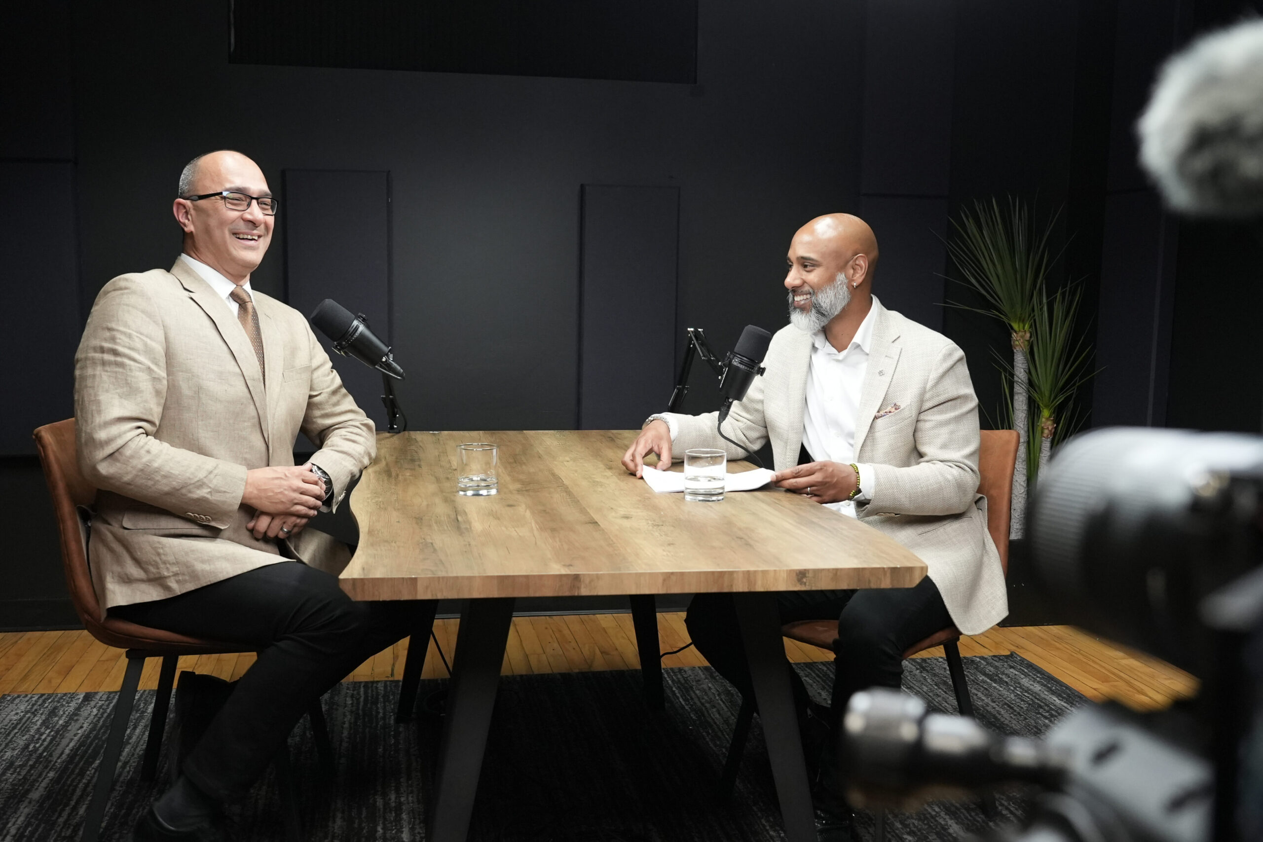 Two men engaged in a podcast discussion at a wooden table, one in a beige suit and glasses, the other in a light gray suit, both smiling with microphones in a modern studio setting.