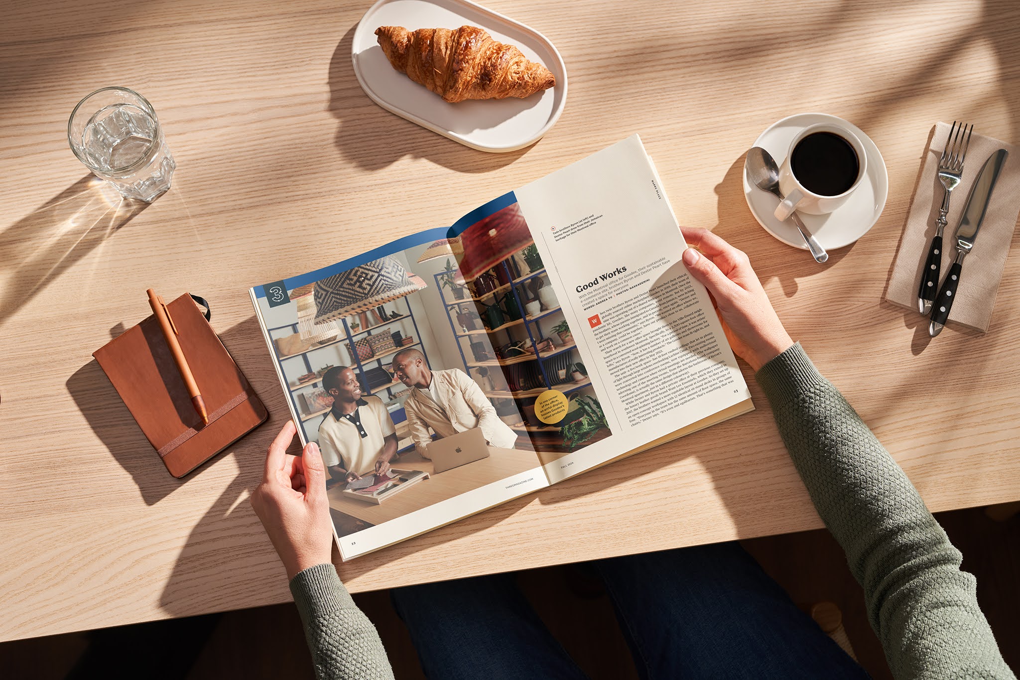 Hands holding an open magazine featuring two men engaged in conversation, with a stylish interior background, accompanied by a croissant, coffee, and notebook on a wooden table, reflecting the theme of storytelling and brand connection in Three Magazine.