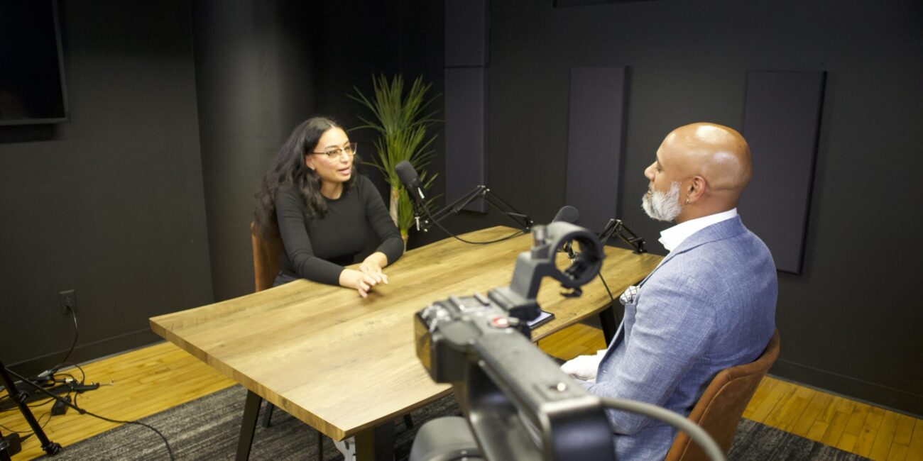 Podcast interview setting with two speakers, one woman in a black top and glasses, and one man in a light blue suit, discussing leadership and cultural identity, with microphones and a camera visible.