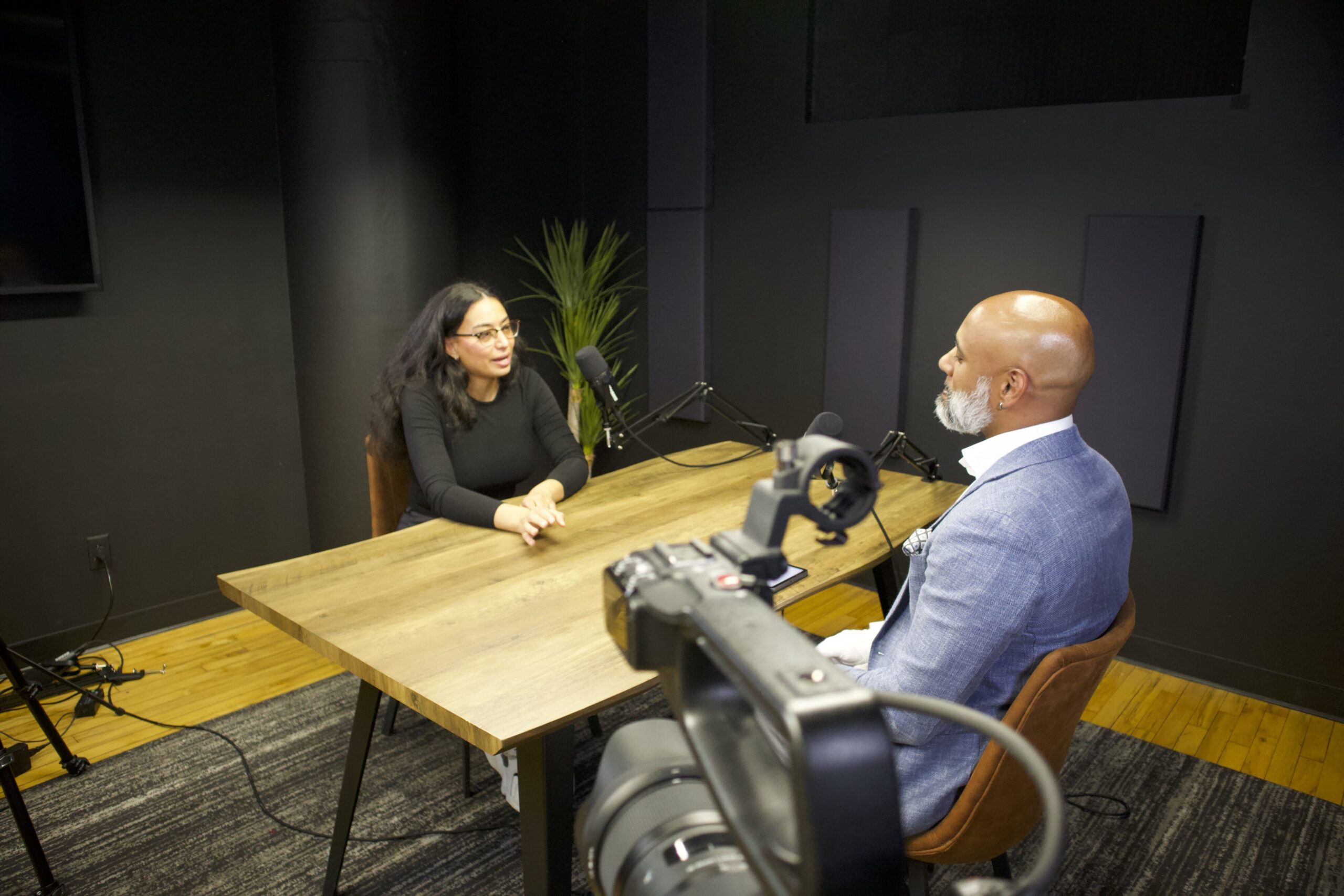 Podcast recording session featuring a woman speaking and a man listening, with microphones and a camera setup in a modern studio environment.