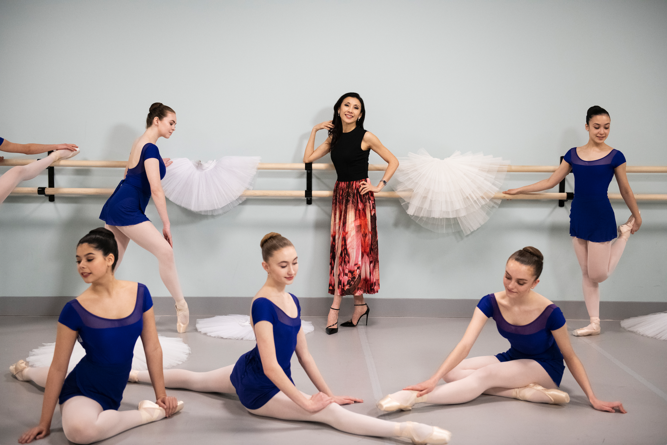 Ballet dancers in blue costumes practicing in a studio, with a female instructor in a colorful skirt, showcasing dance techniques and traditional ballet attire.