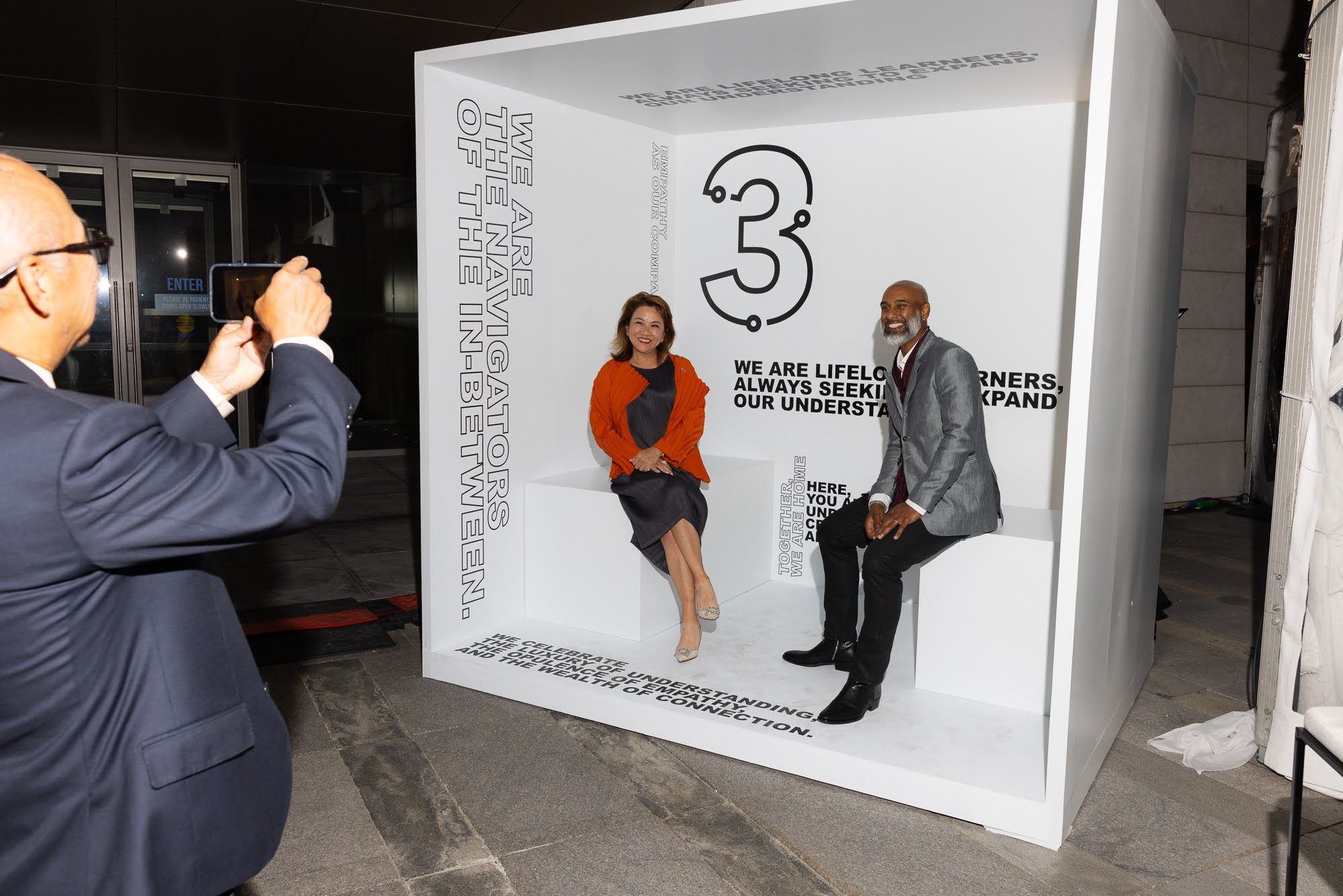 Two diverse individuals seated in a branded white booth with the number "3" and text emphasizing navigation and understanding, while a third person captures the moment with a smartphone, highlighting Three Magazine's theme of connection and storytelling.