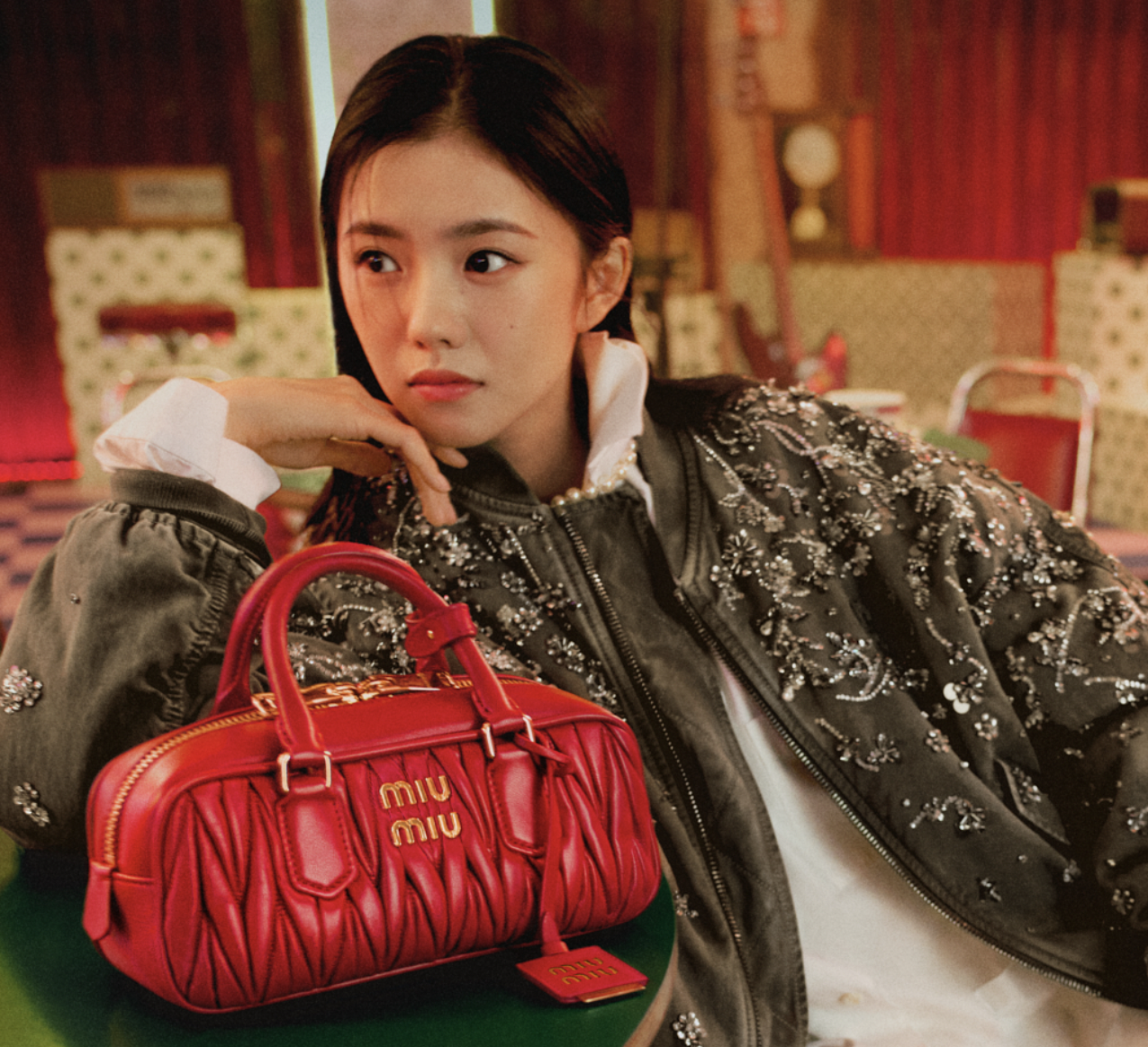 Woman in embellished bomber jacket resting her chin on hand beside a red Miu Miu handbag in a stylish, retro-inspired setting.