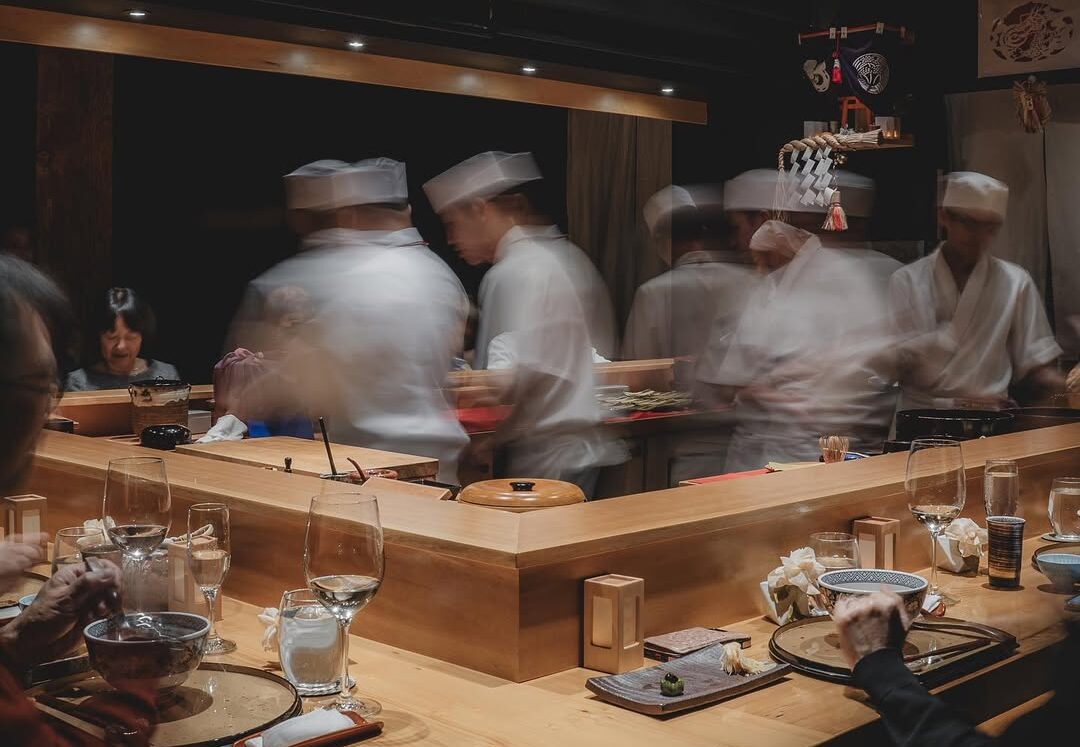 Sushi chefs in motion preparing dishes at a traditional Japanese restaurant, showcasing a vibrant culinary atmosphere with patrons enjoying their meals.