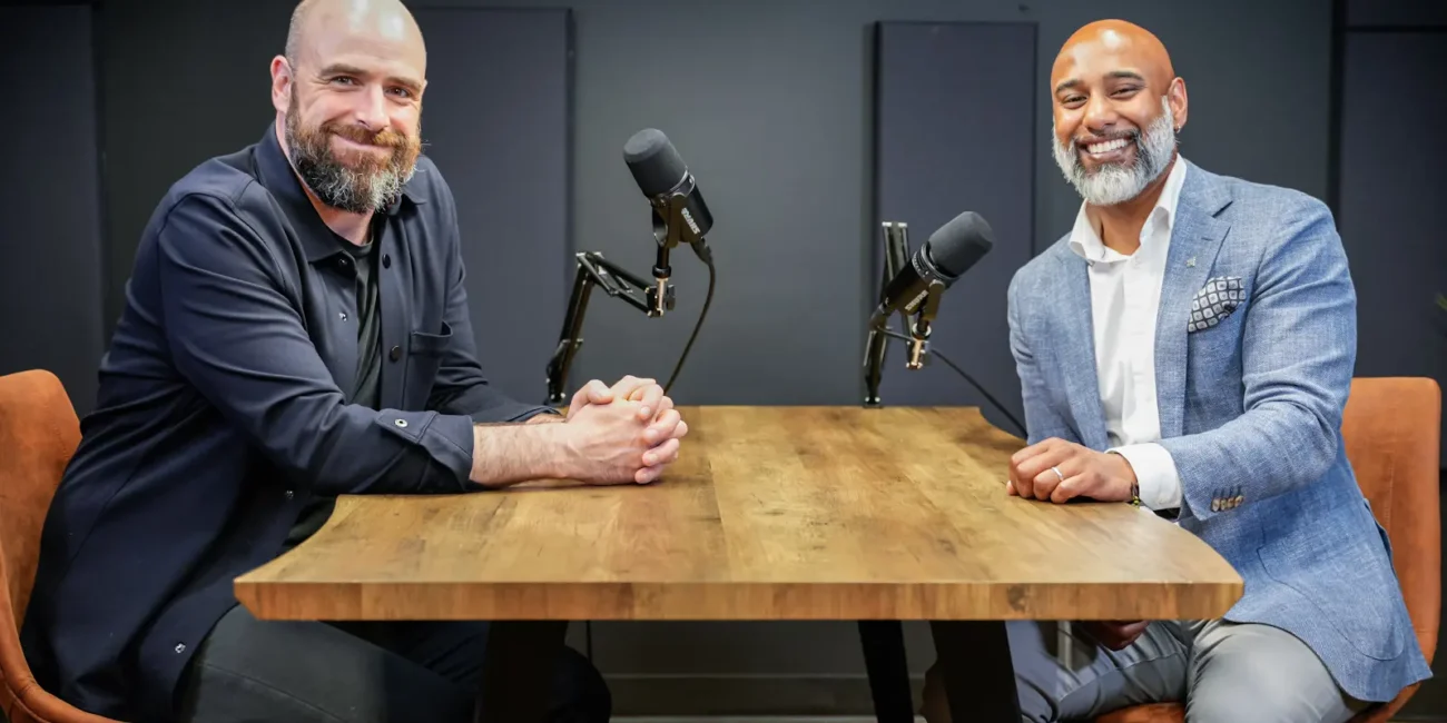 Muraly and Daniel seated at a podcast table, discussing leadership and culture, with microphones in a modern studio setting.