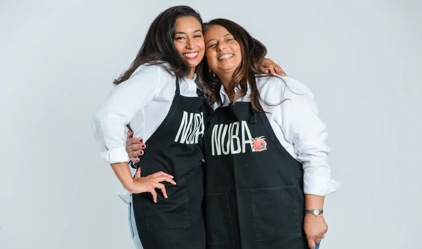 Amal Soliman and Gina El Kattan, co-founders of Nuba Tisane, smiling together in matching black aprons featuring the Nuba logo, celebrating their Egyptian heritage and homemade hibiscus tea.