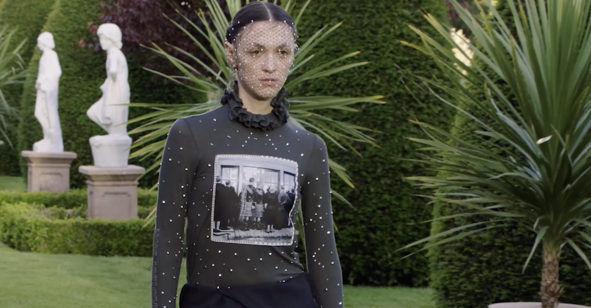 Model wearing a black outfit with a historical photograph print, adorned with sequins, in a lush garden setting with statues and greenery, reflecting themes of fashion and identity.