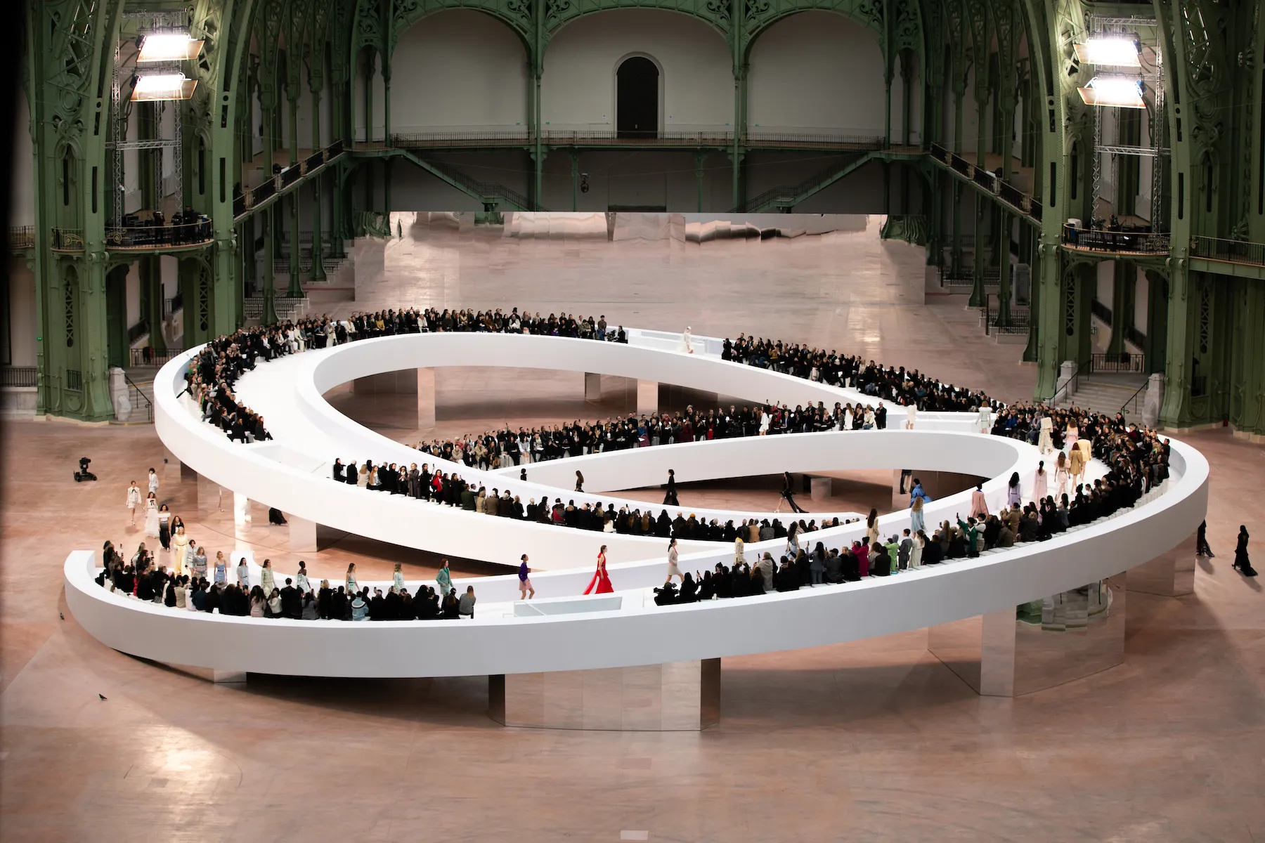 Fashion show featuring models walking on a unique spiral runway in a grand venue, surrounded by an audience of stylish attendees, highlighting luxury fashion and contemporary design.
