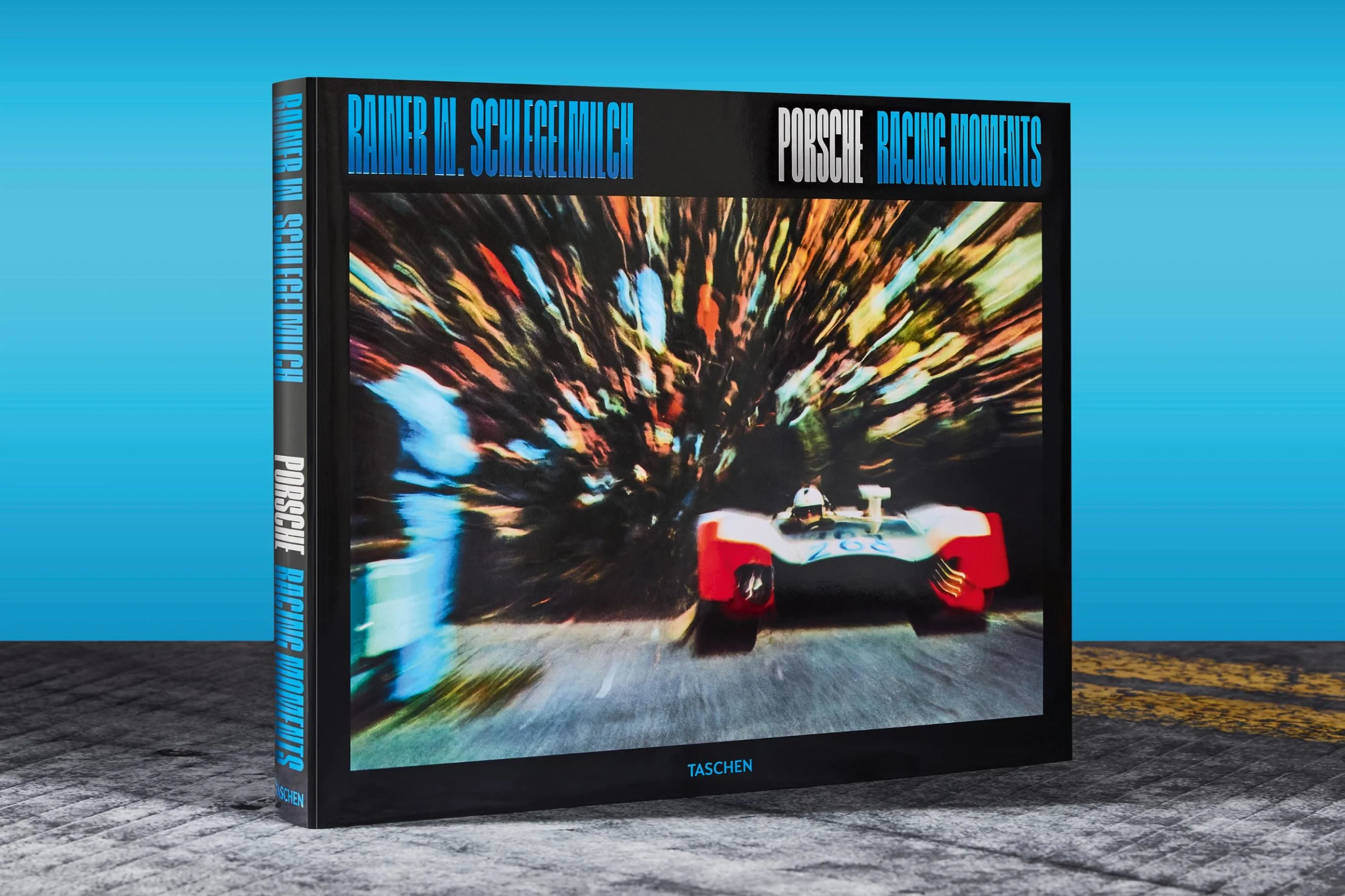 Book titled "Porsche Racing Moments" by Rainer W. Schlegelmilch, featuring a blurred image of a racing car, symbolizing speed and motion, against a vibrant blue background, emphasizing the dynamic nature of automotive art and culture.