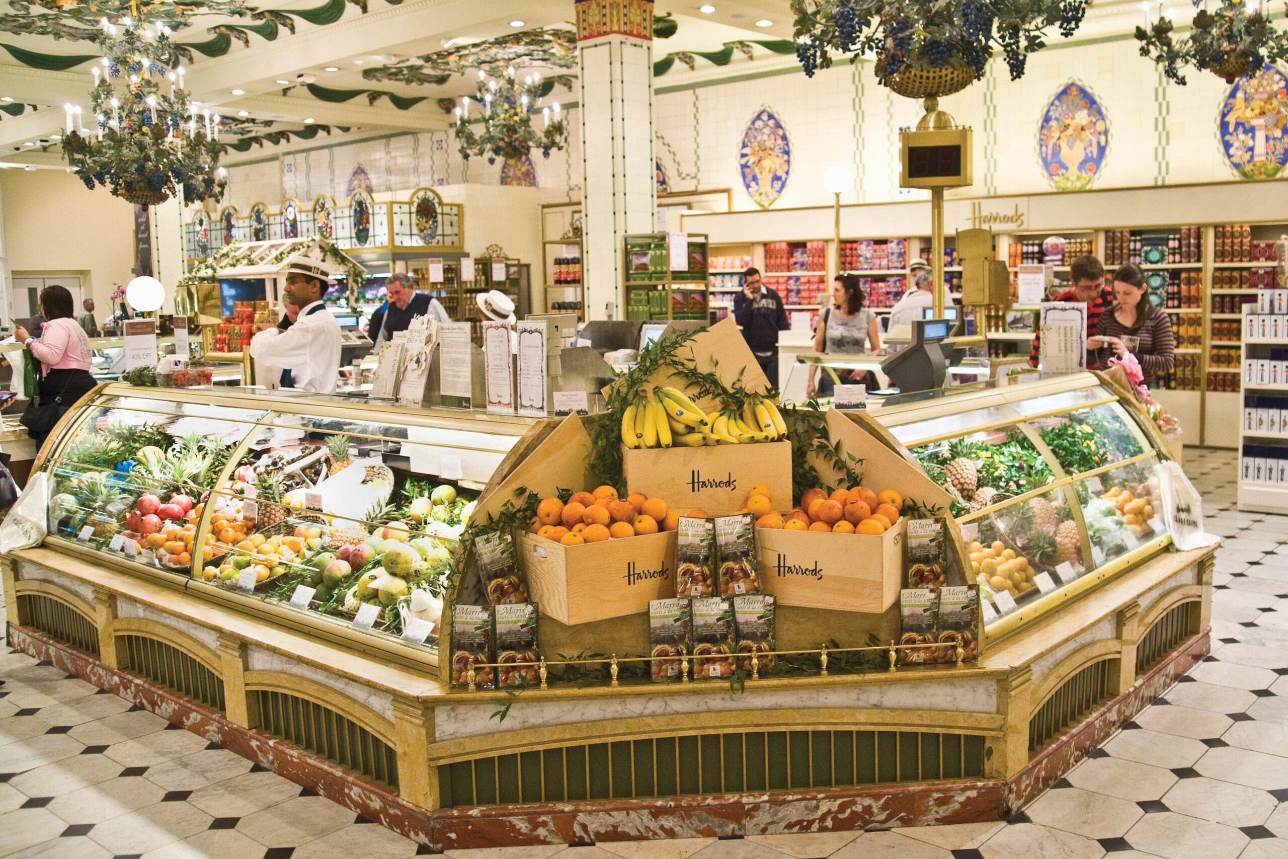 Gourmet grocery display at Harrods featuring fresh fruits, including oranges and bananas, with shoppers in a luxurious market setting.