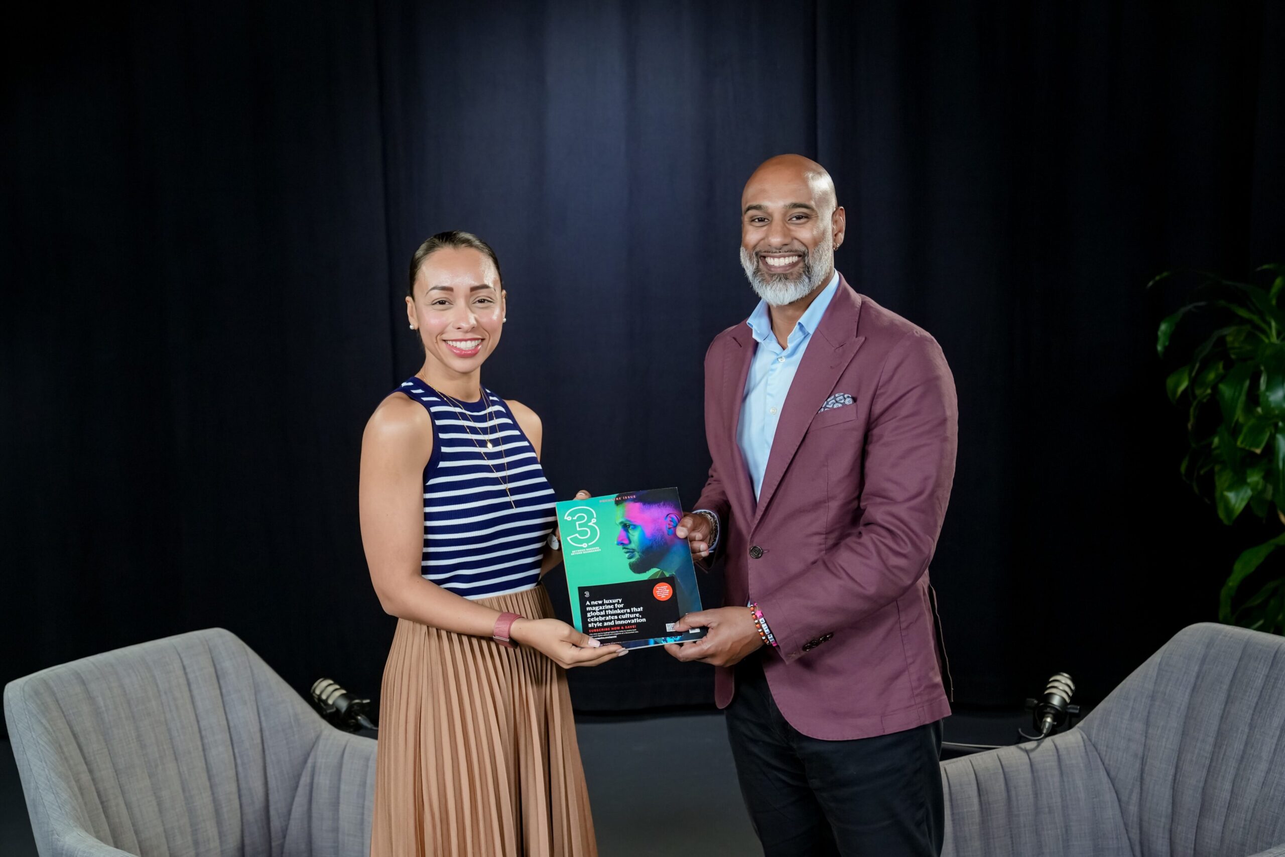 Two individuals smiling and holding a magazine featuring the number "3," set against a dark background, with a plant and seating visible, reflecting themes of inclusivity and cultural diversity in professional settings.