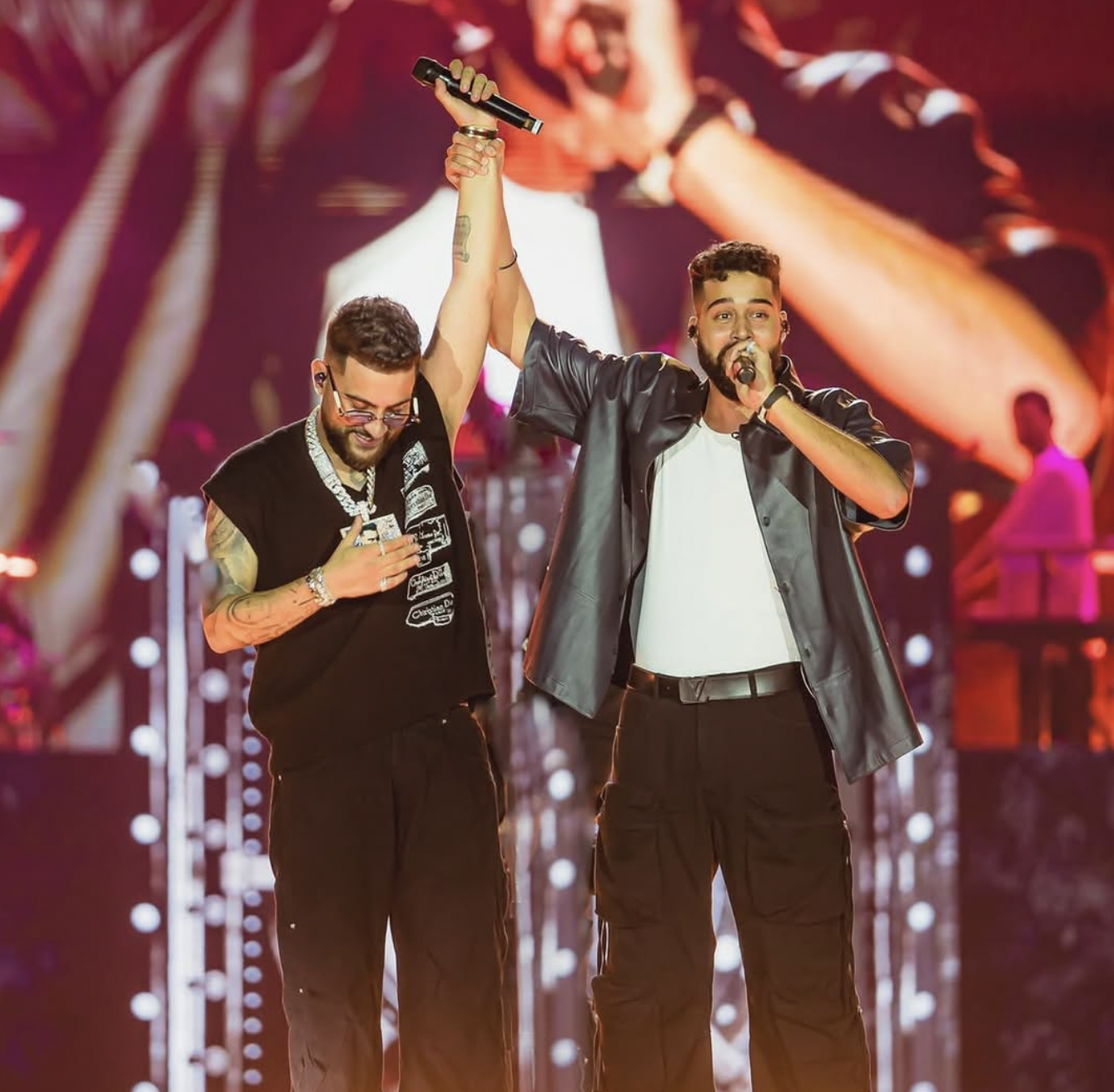 Two male musicians performing on stage, one holding a microphone and raising the other's hand in celebration, with a vibrant concert backdrop.