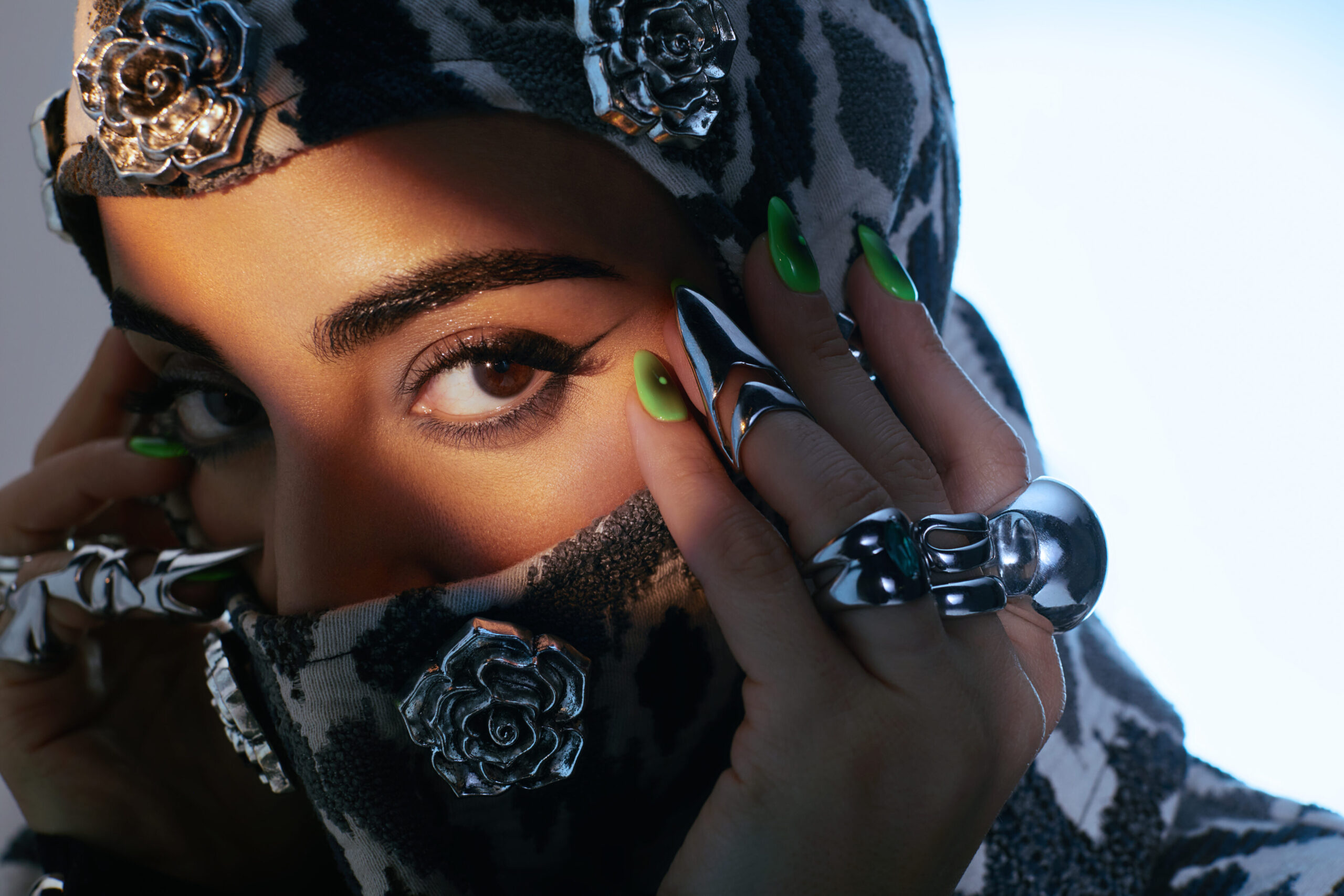 Fashion model with striking green nails and silver jewellery, wearing a patterned headscarf adorned with floral embellishments, showcasing a bold and innovative style.