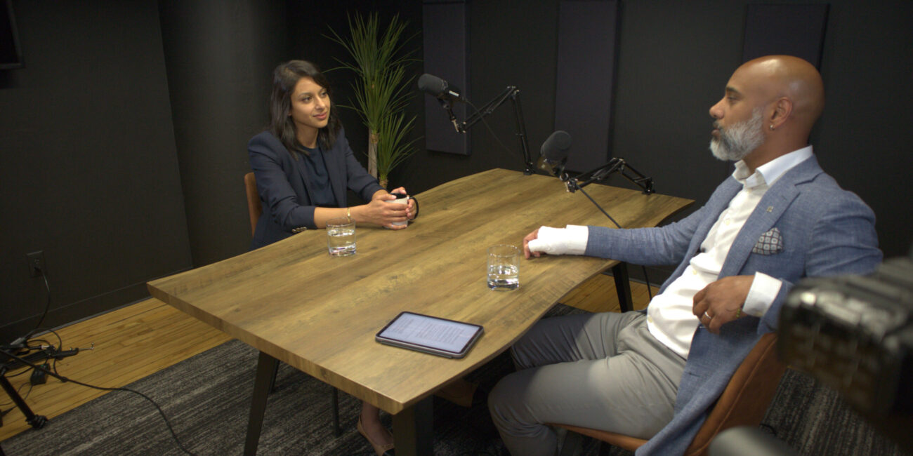 Neha Khera and a podcast host engaged in conversation at a table, discussing investment and entrepreneurship in a modern studio setting.