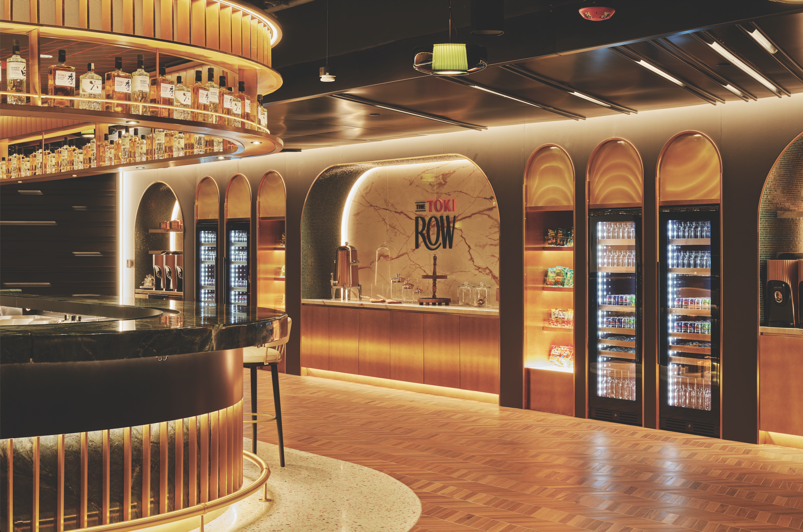 Modern bar interior featuring a sleek black marble countertop, illuminated shelves displaying various liquor bottles, and a stylish backdrop with "THE TOKI ROW" signage, complemented by warm lighting and elegant wooden flooring.