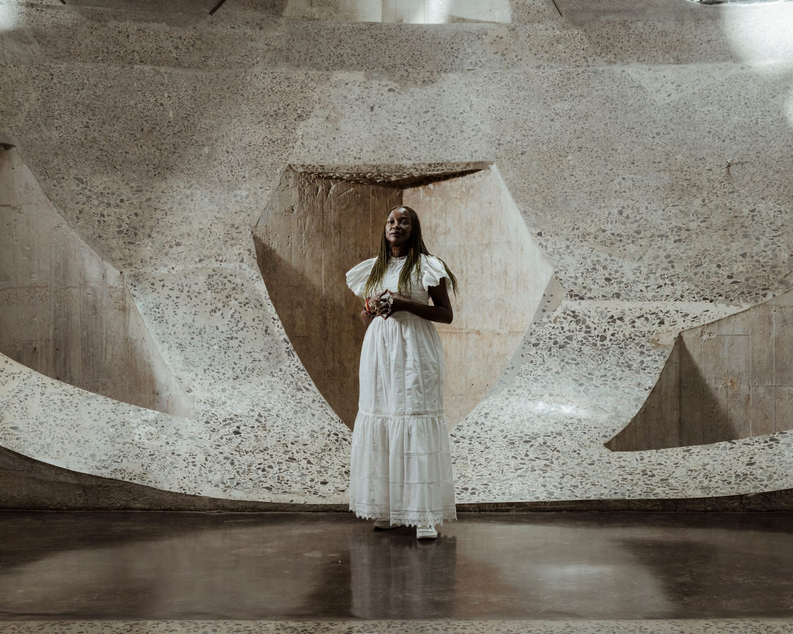 Alyssa Alikpala standing in an art installation at the Porsche SCOPES festival in Toronto, wearing a flowing white dress, surrounded by textured concrete walls showcasing modern art.
