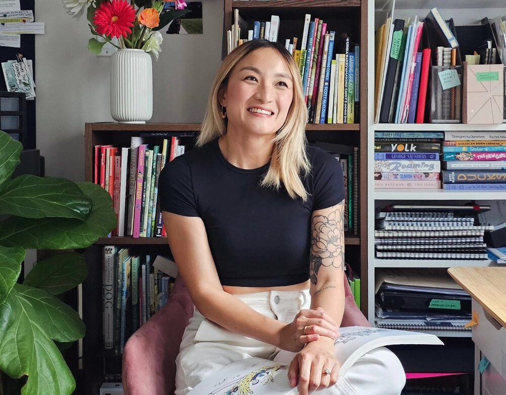 Artist Zoe Si smiling while seated in a cozy workspace surrounded by colorful books and plants, reflecting the creative spirit of noctourism and nighttime experiences.