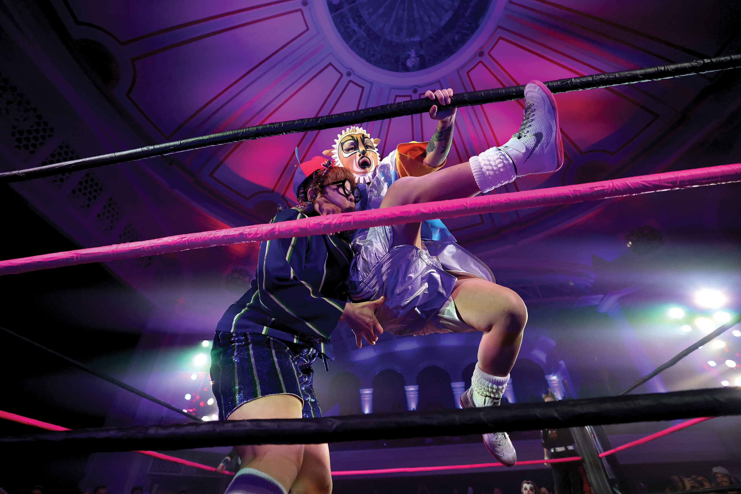All-women's wrestling match in a vibrant arena, featuring a wrestler in a clown-inspired costume and another in athletic gear, engaged in a dynamic move within the ring, surrounded by colorful lights and an enthusiastic crowd.