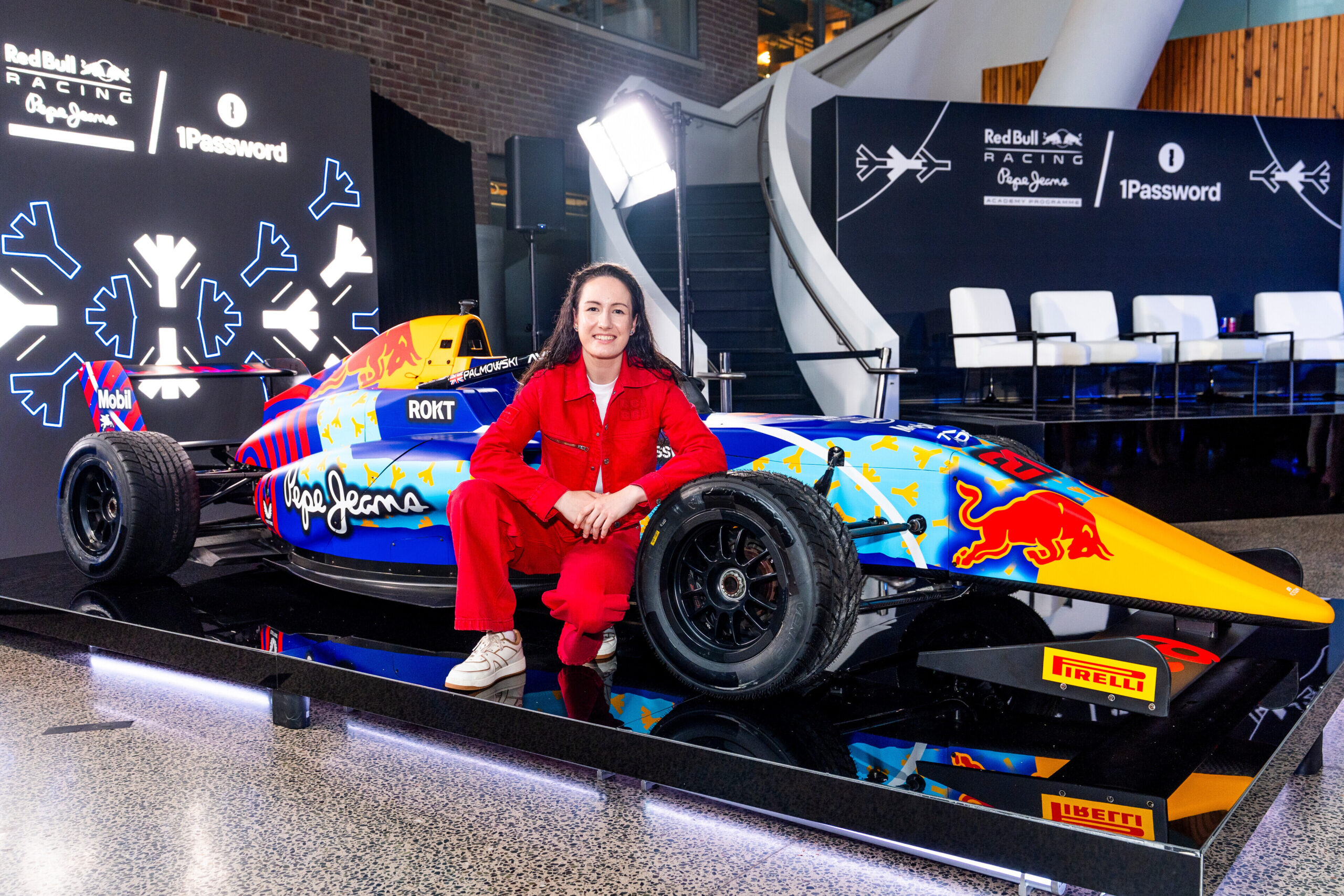 Female race car driver seated beside a colorful Red Bull Racing car, showcasing a blend of technology and motorsport culture in a modern event space.