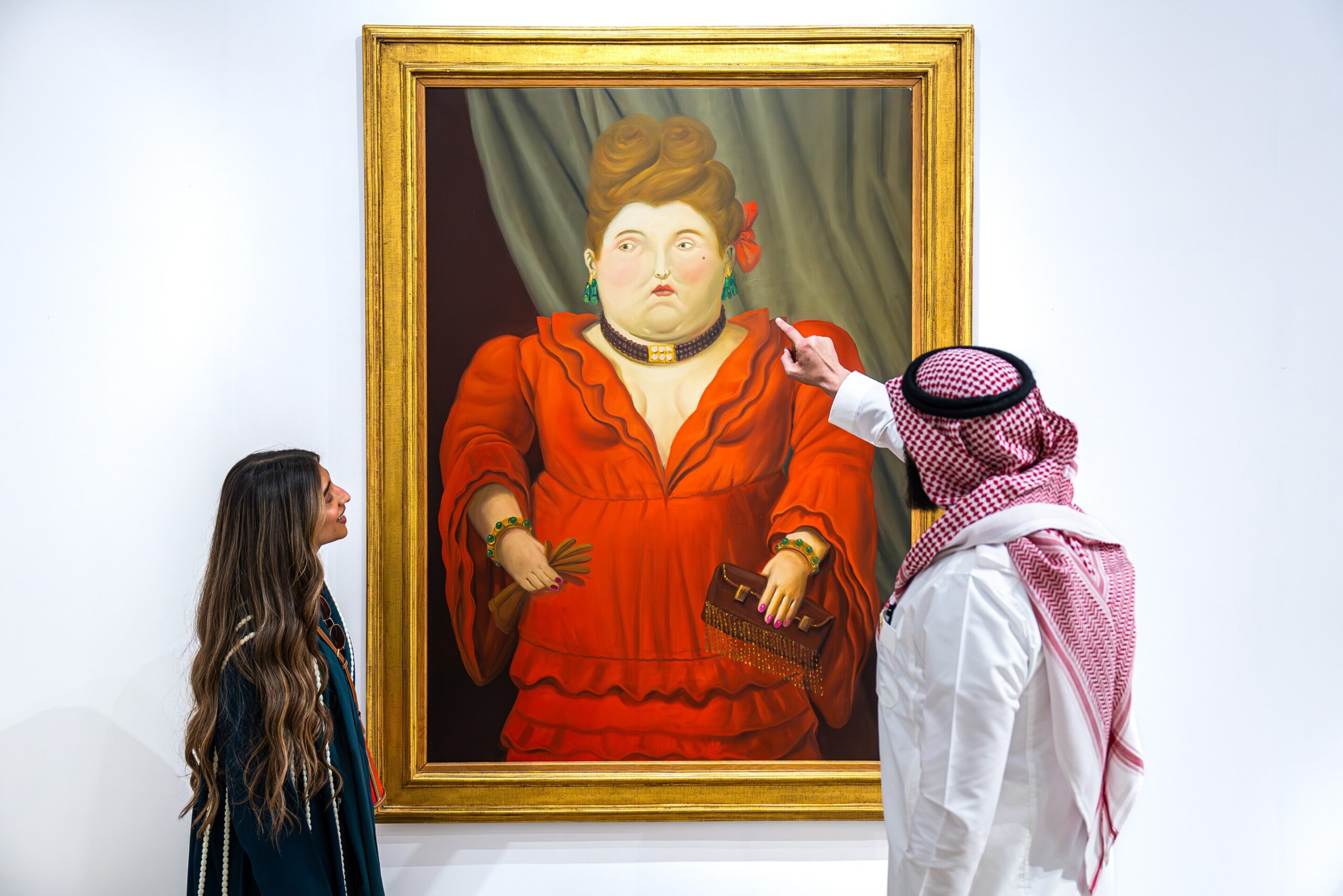 Woman admiring a large painting of a woman in a red dress, while a man in traditional attire points at the artwork, highlighting cultural engagement in an art gallery setting.