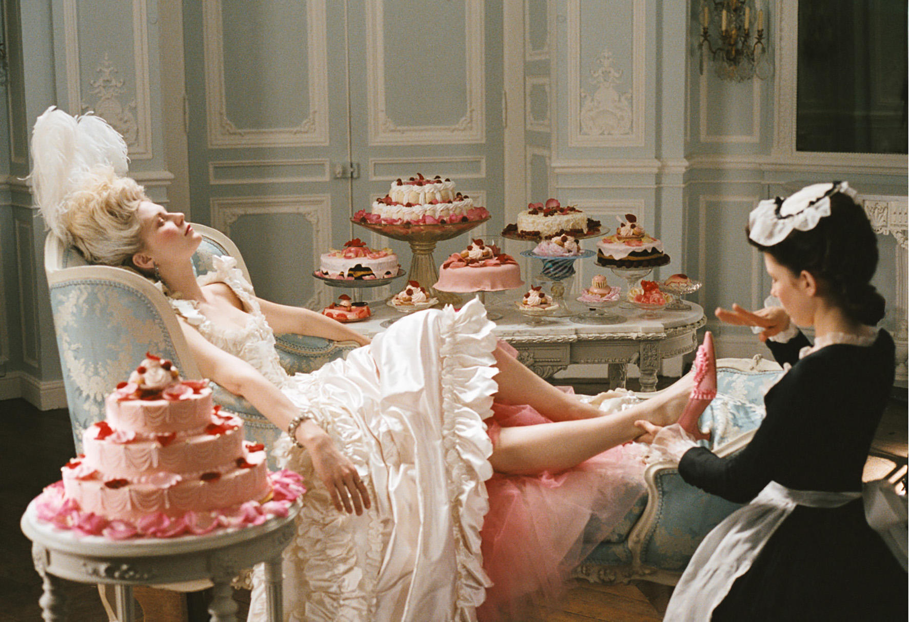 Woman in elegant dress reclining on a chaise lounge, surrounded by an array of decorative cakes and pastries, with a maid attending to her feet in a lavish interior setting.