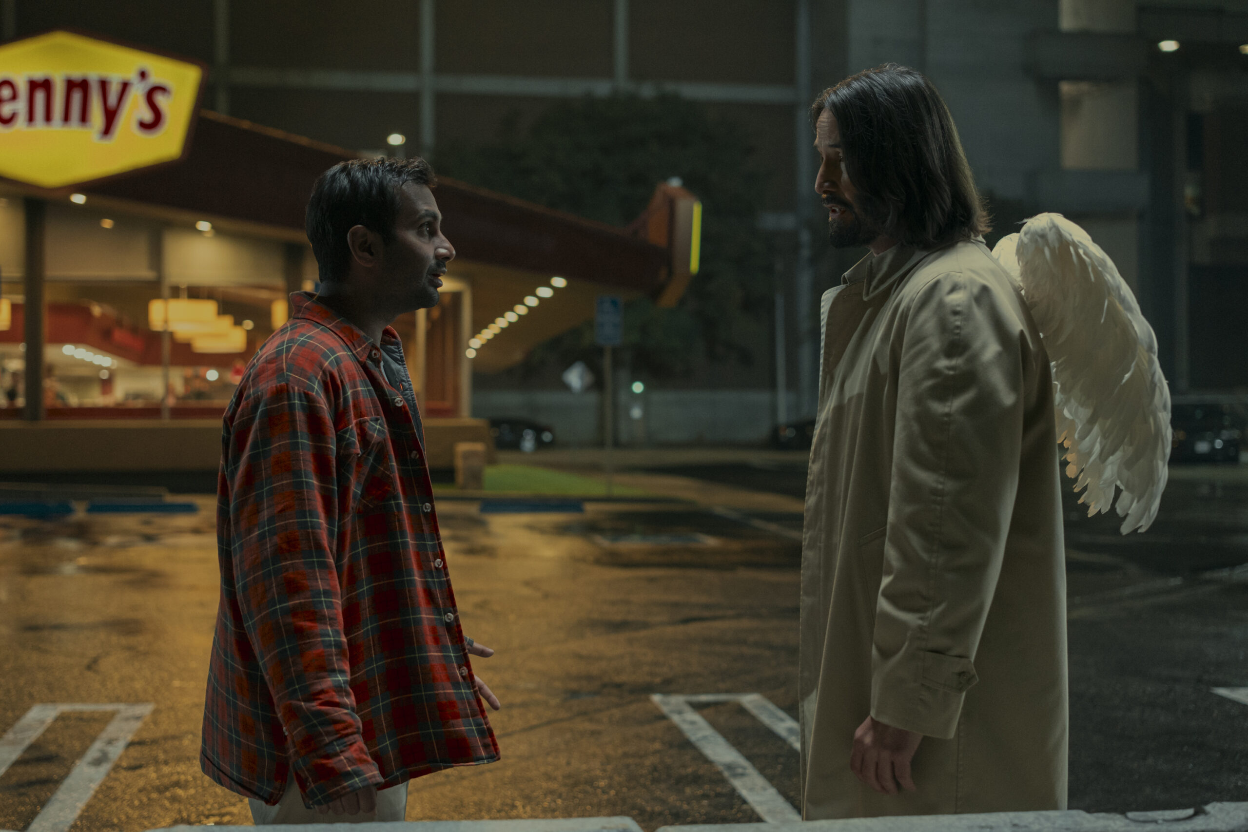 Two men facing each other in a parking lot at night; one wears a plaid shirt and the other a long beige coat with angel wings, illuminated by the Denny's restaurant sign in the background.