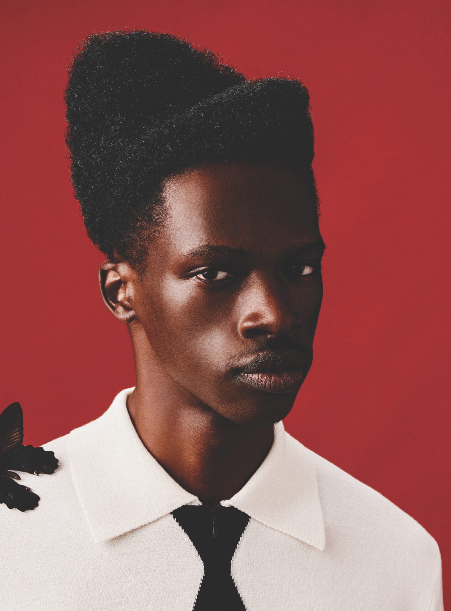 Portrait of a young man with textured hair styled in a bun, wearing a white shirt with a black tie and a butterfly detail, set against a vibrant red background, reflecting contemporary fashion aesthetics.